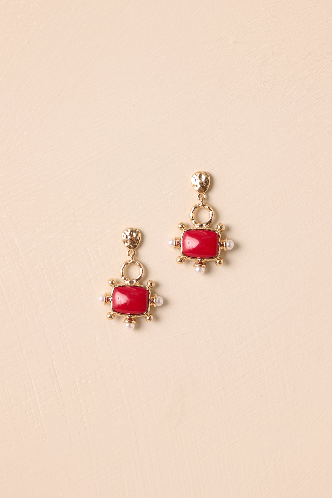 Gold drop earrings displayed against beige background featuring hammered studs, open circles, and red resin squares.