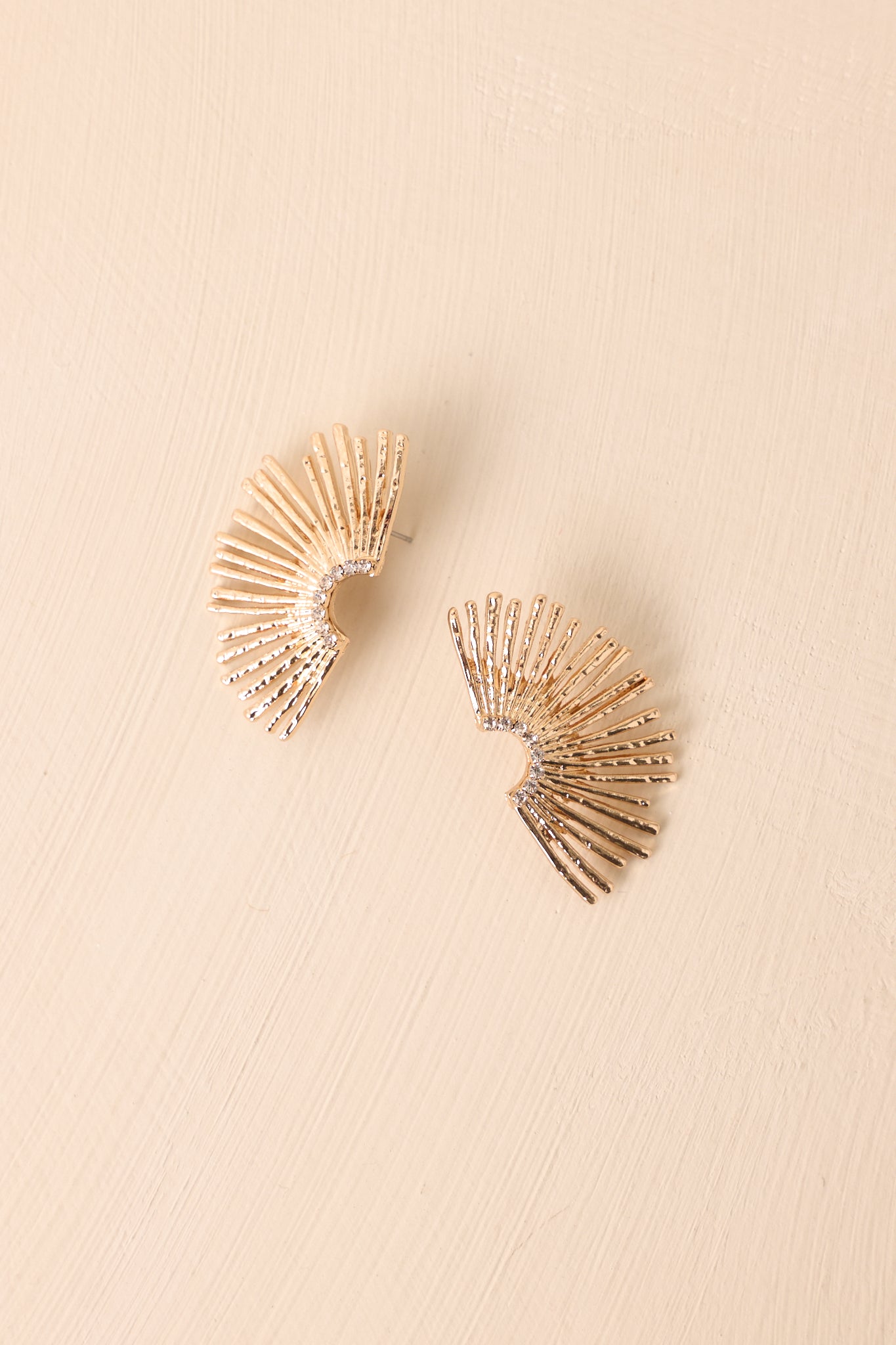 Gold sunburst fan earrings displayed on a beige background, showing the rhinestone detailing along the curved center.