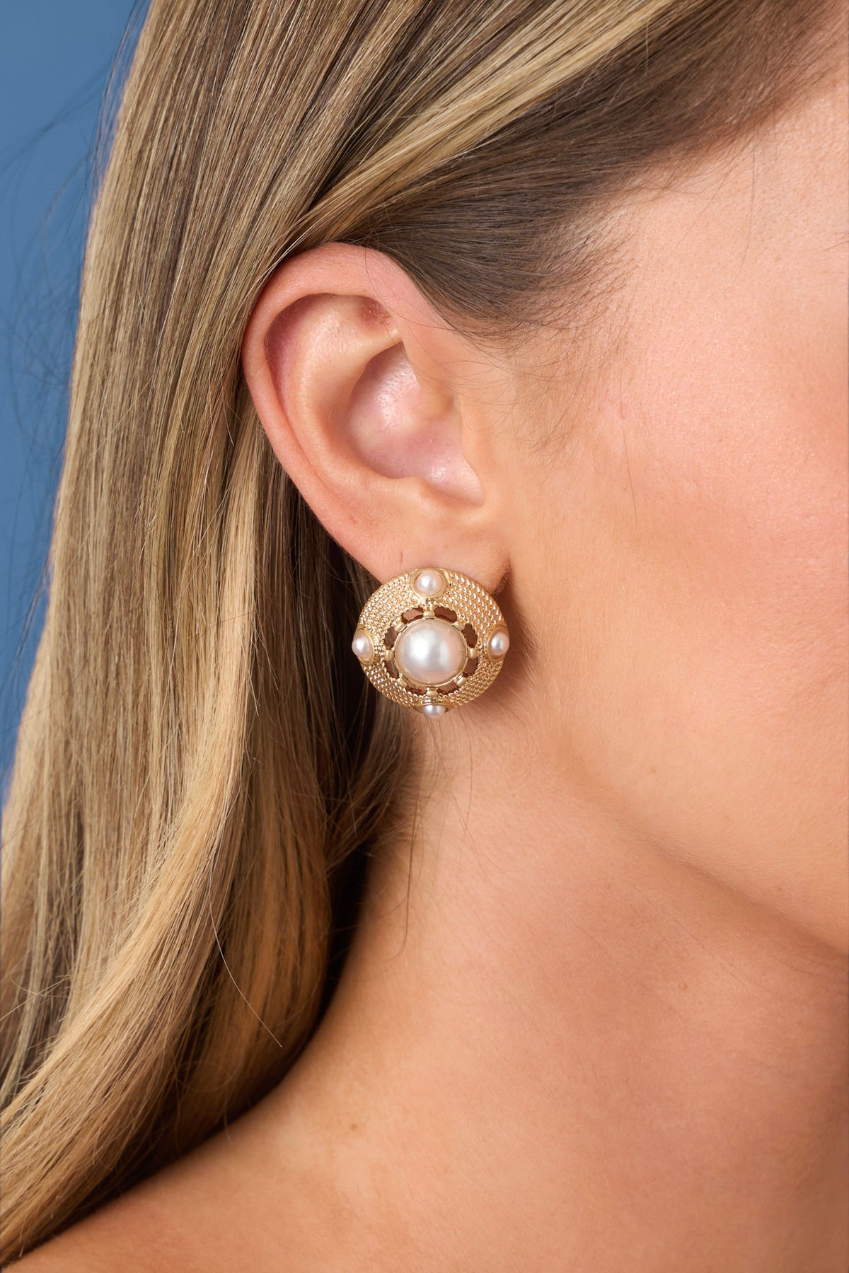 Circular pearl stud surrounded by gold hardware, against a blue backdrop. 