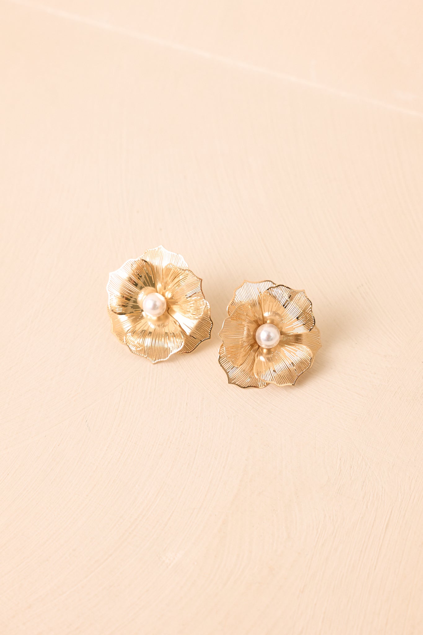 Gold floral earrings displayed against a beige background, highlighting their dimensional petals and luminous faux pearls.
