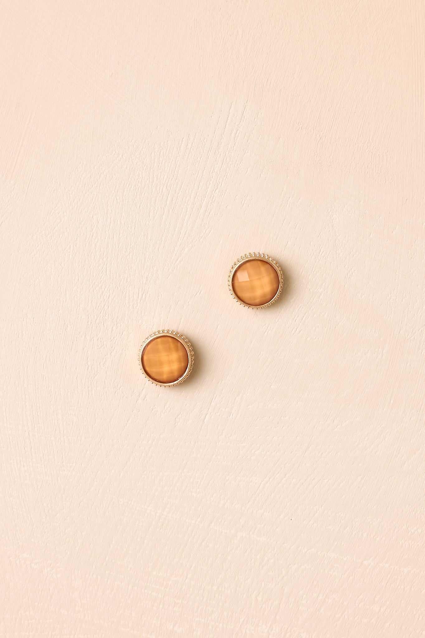 Gold halo stud earrings displayed against a beige background, highlighting the vibrant orange stone and delicate setting.
