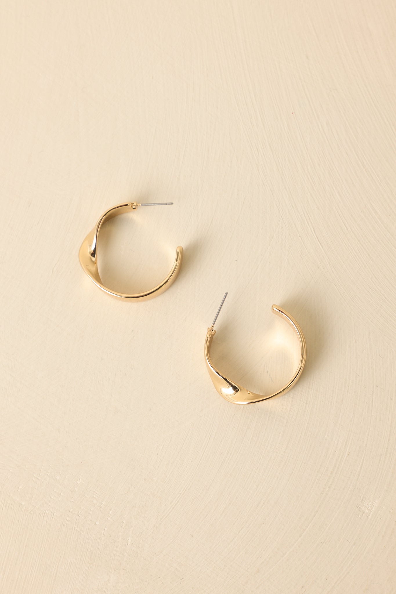 Gold hoop earrings displayed on a beige background, showcasing the flat band design and subtle front twist.