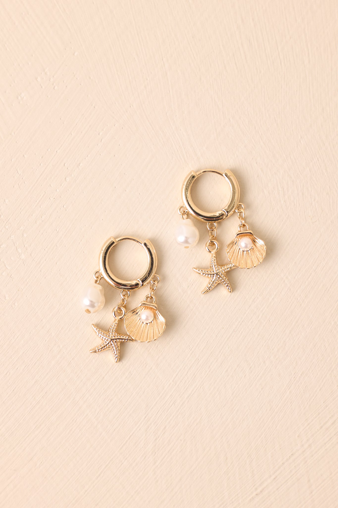 Gold huggie earrings with ivory pearls and whimsical ocean-themed charms against a beige backdrop.