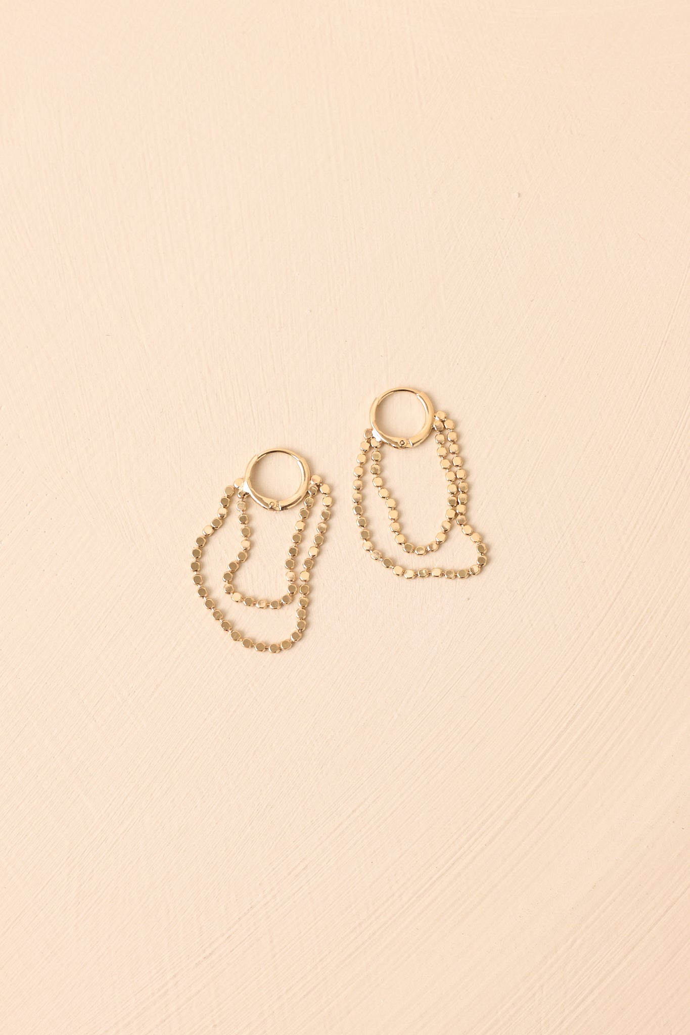 Gold huggie hoop earrings displayed against a beige background, showing the two disc drop chains and polished surface.