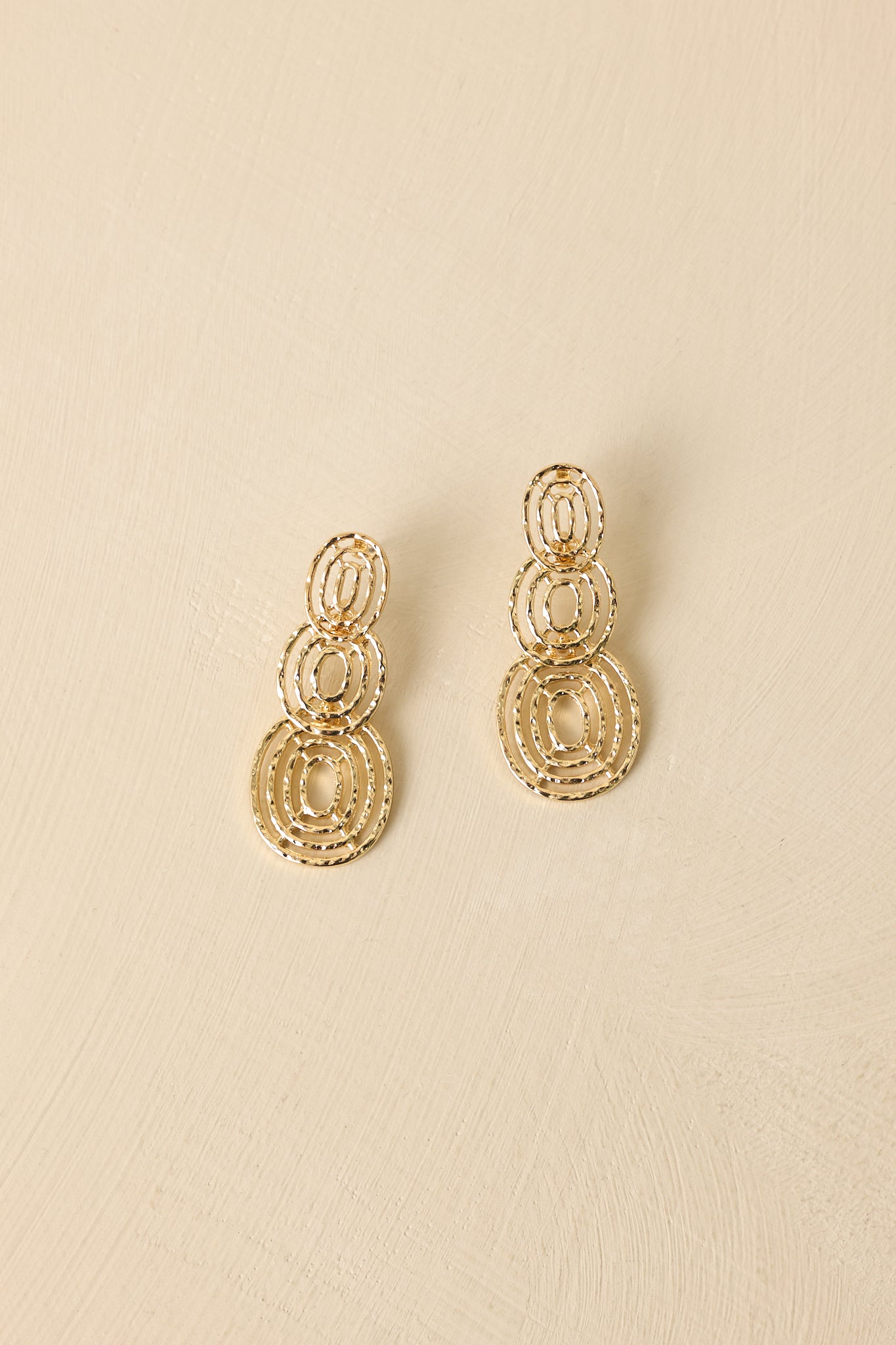 Gold layered circle earrings displayed against a beige background featuring open ring details and warm shine.