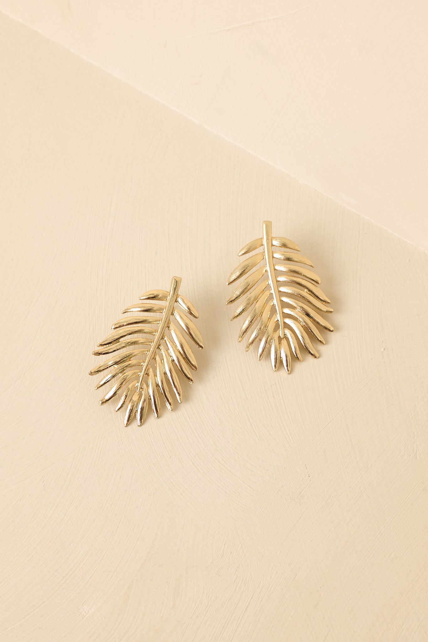 Front view of gold palm leaf earrings displayed on beige background emphasizing elongated shape.