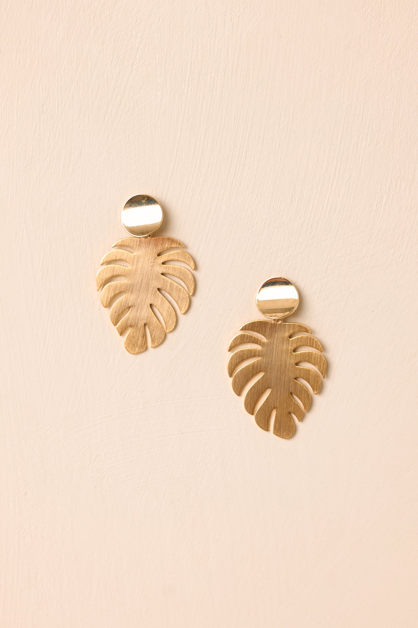Gold palm leaf earrings displayed against a beige background featuring polished discs and brushed textures.