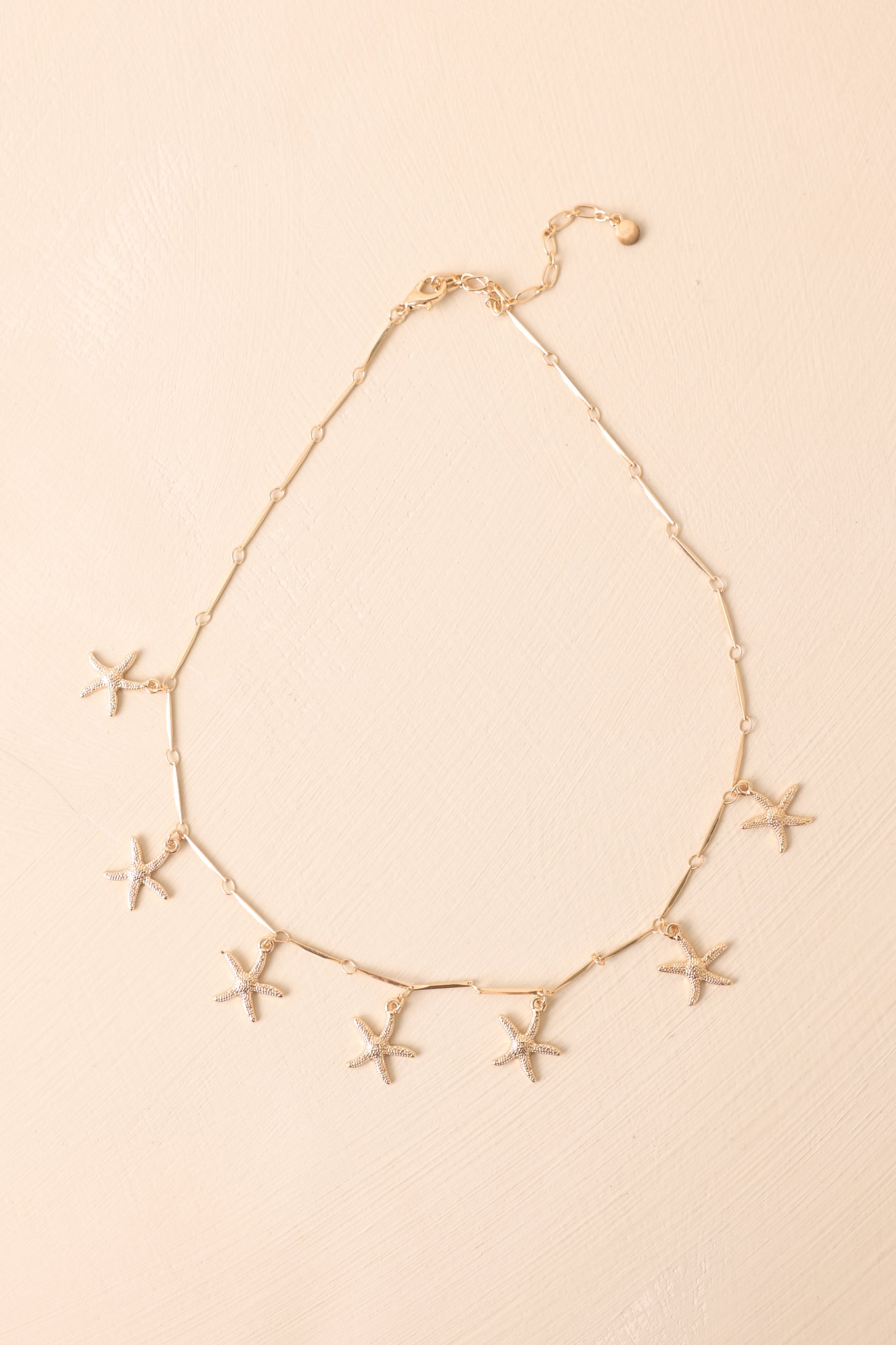A complete shot of the gold paper clip chain necklace displayed on a beige background featuring multiple textured starfish charms.