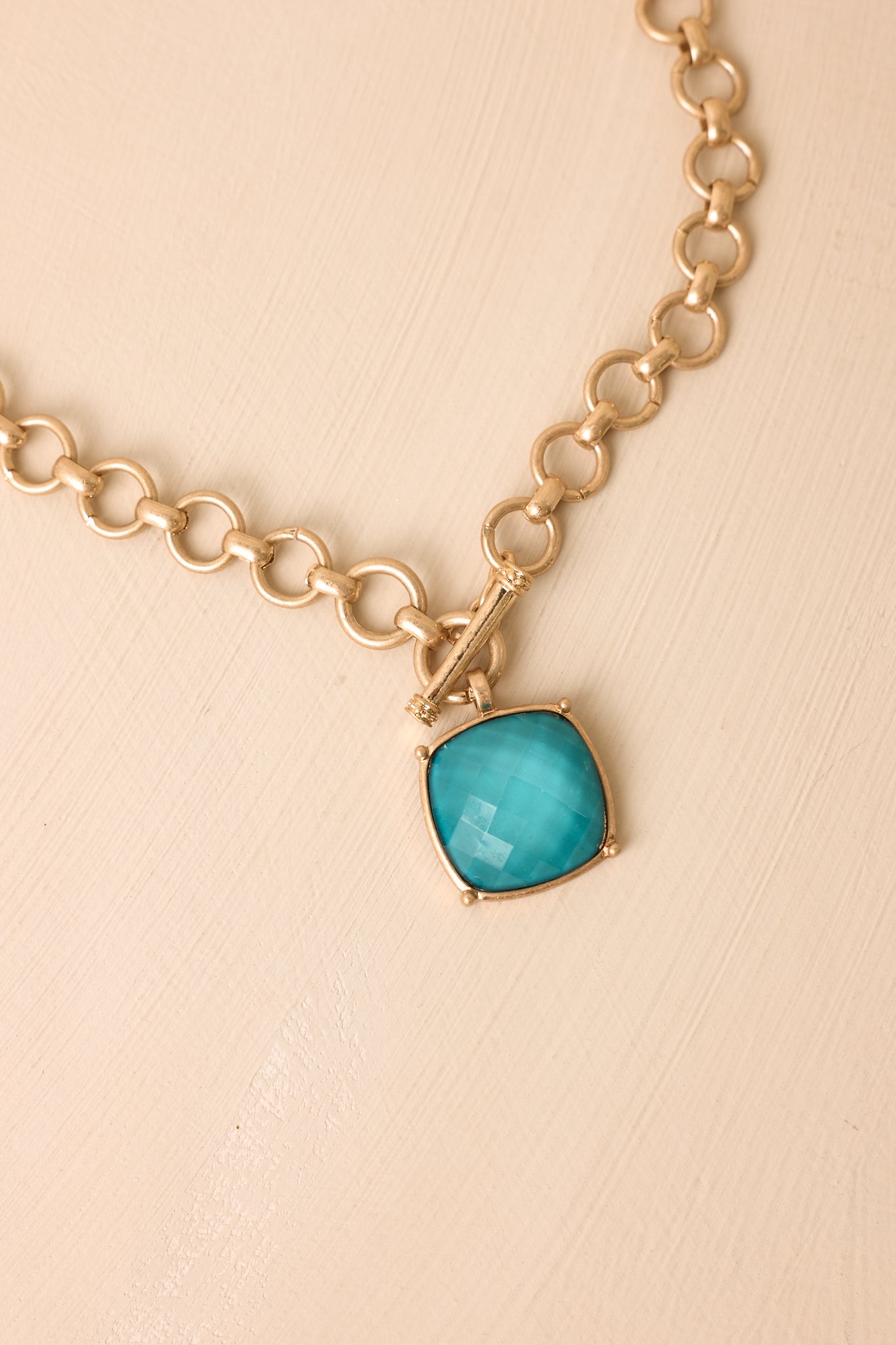 Matte gold necklace displayed with a faceted teal pendant and modern front toggle clasp.