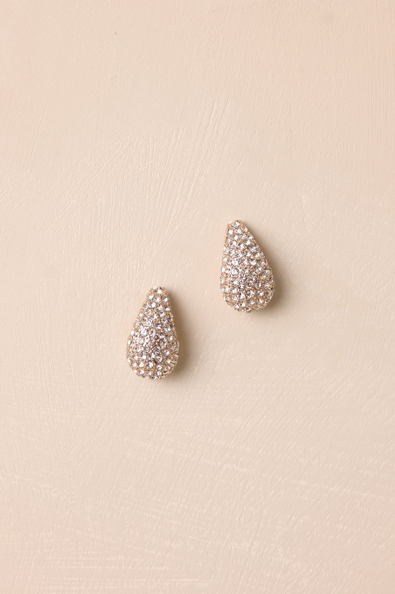 Gold rhinestone teardrop earrings displayed against a beige background featuring a dimensional silhouette and shimmering finish.