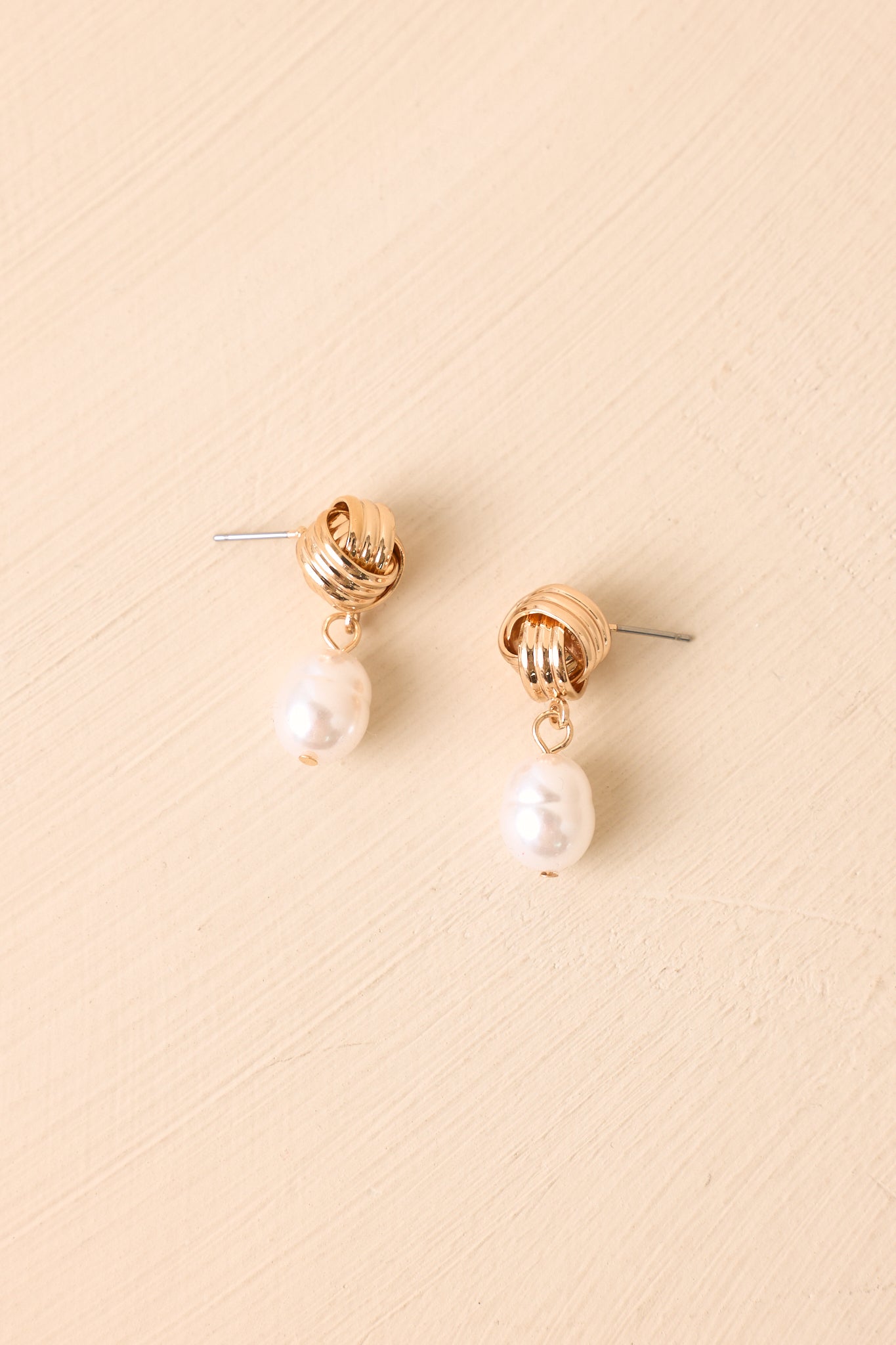 Gold-tone earrings displayed against a beige background highlighting the intricate knot design and pearl accents.