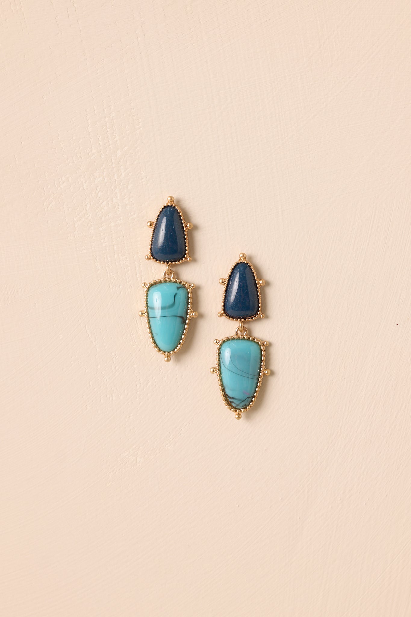 Gold-tone earrings featuring a dark navy faux stone above a larger turquoise stone on beige background.