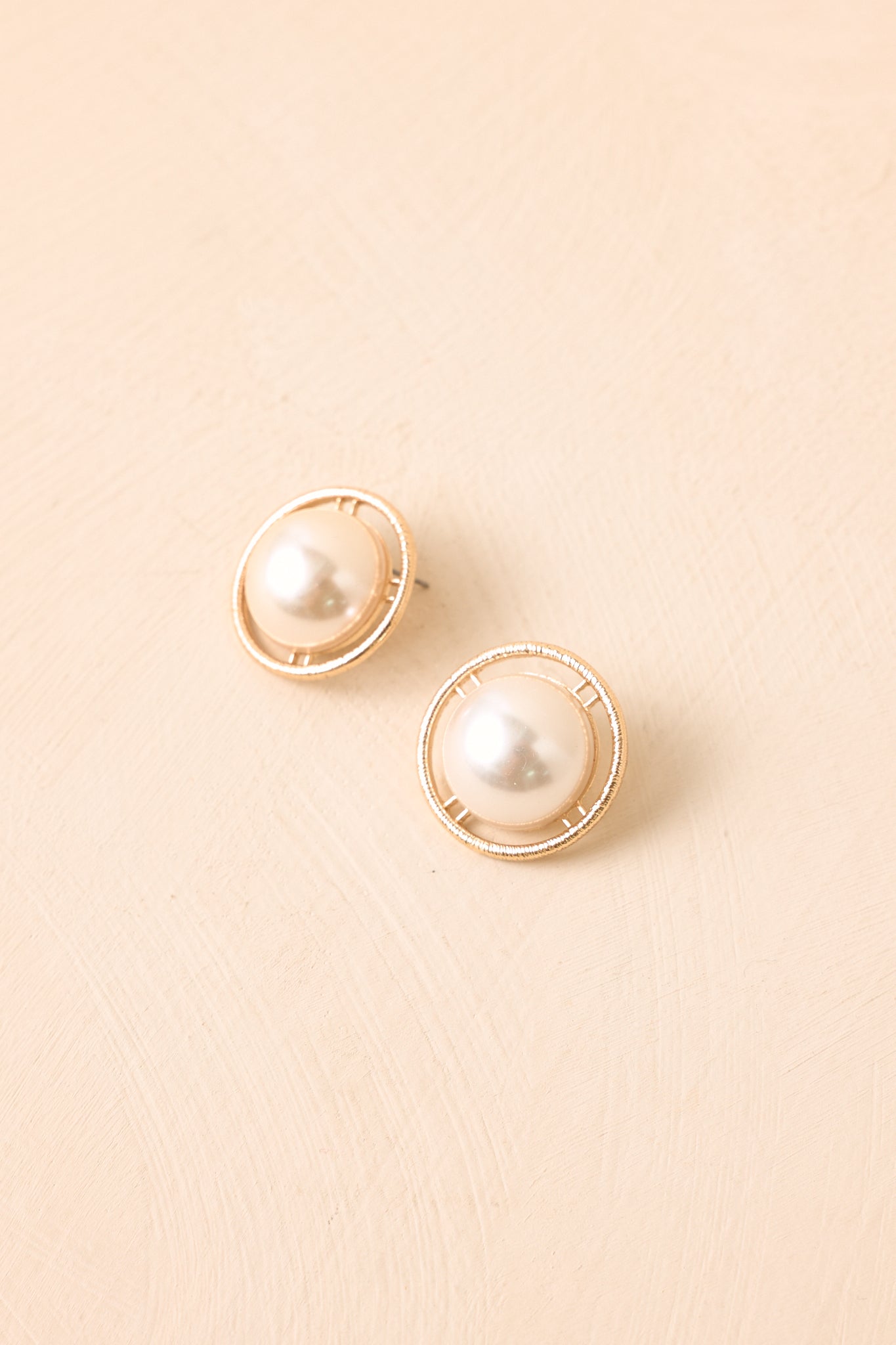 Gold-tone earrings with elegant pearl centers and vintage-inspired texture presented on a neutral beige backdrop.