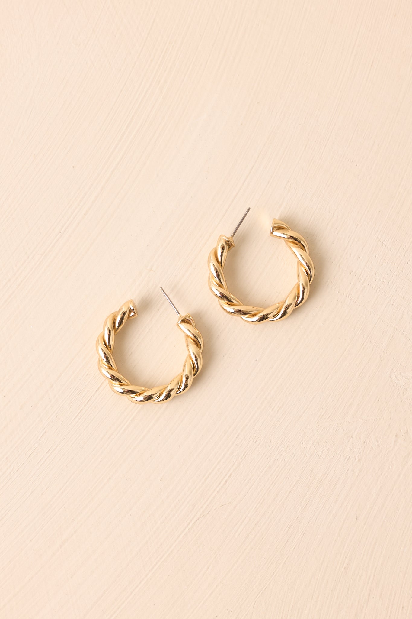 Gold twisted hoop earrings displayed on a beige background, emphasizing the open C-shape and sculptural twist.