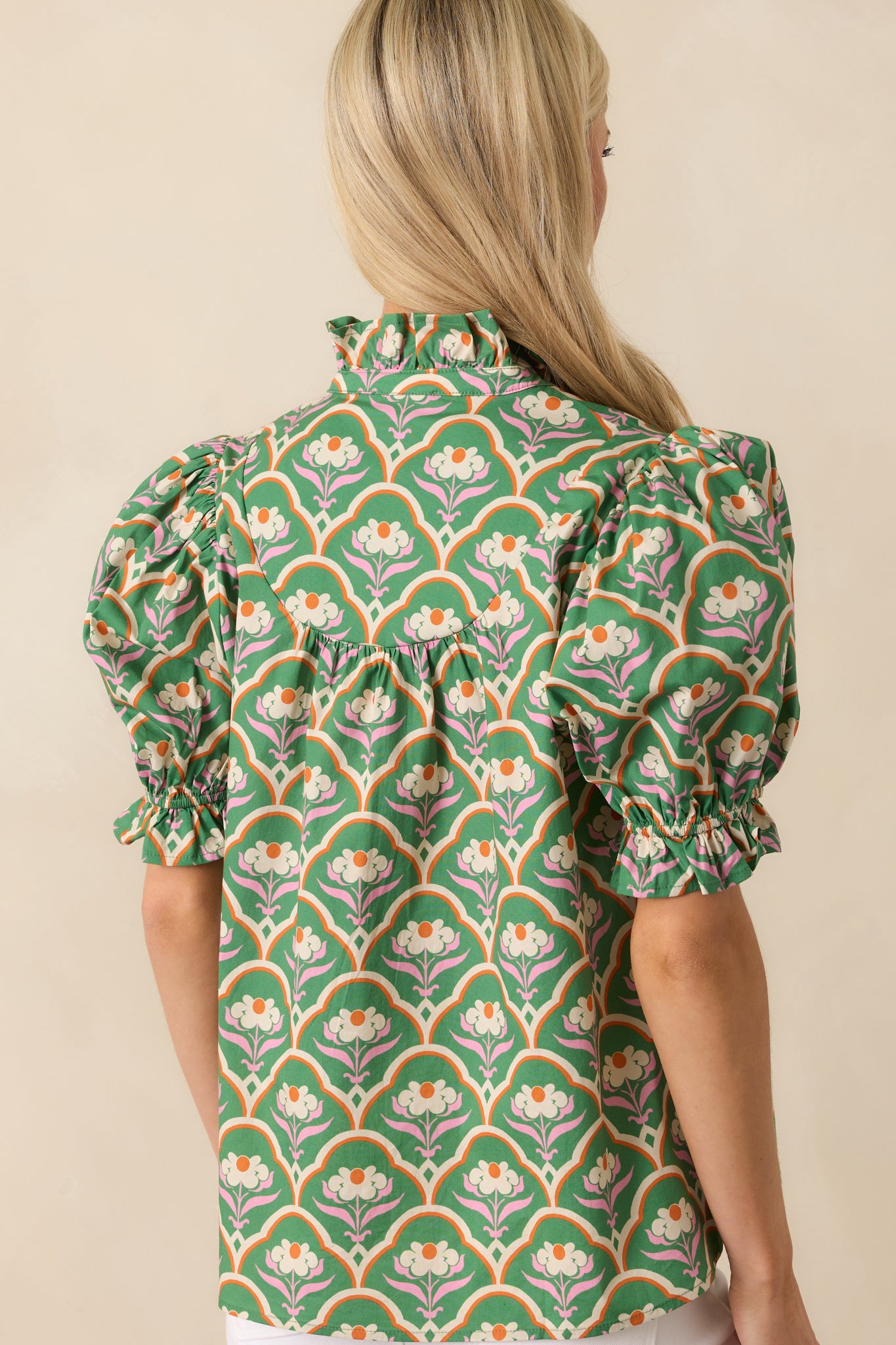 Up close photo of model showing back wearing green top that features a pink multicolor geometric floral print, ruffle notch v-neckline, functional button front, short puff sleeves, ruffle elastic cuffs, and a relaxed fit.