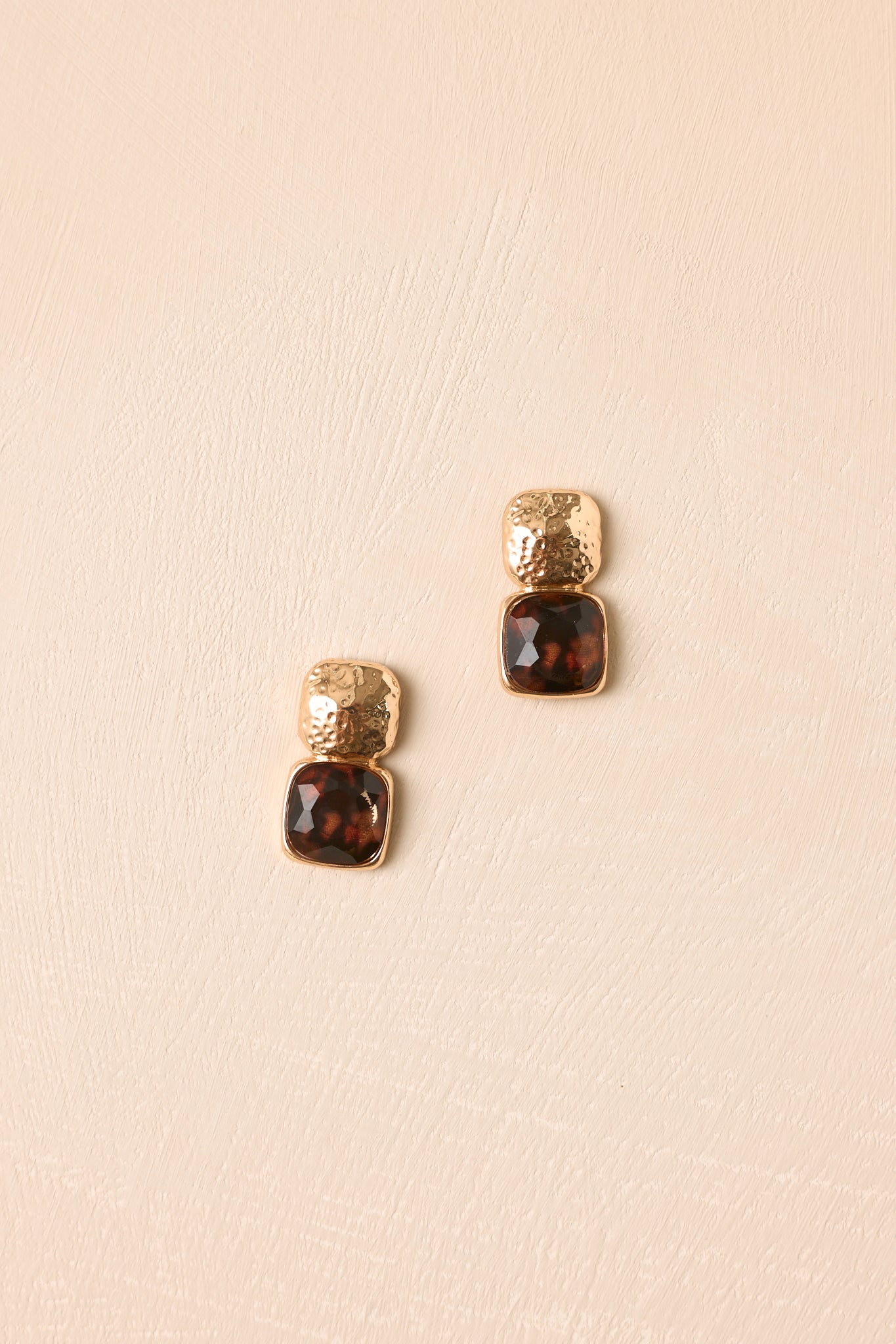 Gold-tone earrings displayed against a beige background, featuring hammered stud tops and glossy tortoise acetate drops with a rounded square silhouette.