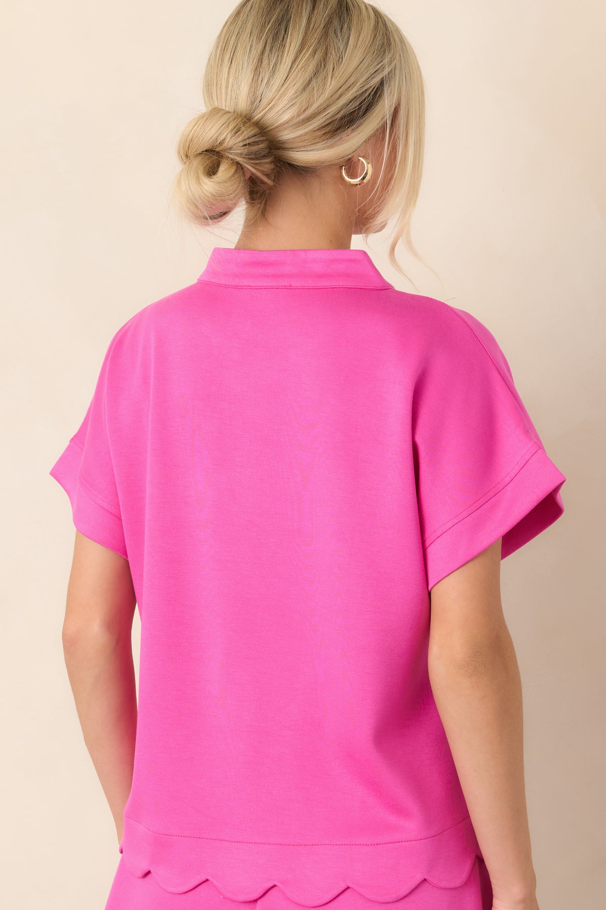 Rear view of the hot pink top, highlighting its relaxed silhouette, short cuffed sleeves, structured fabric, and scalloped hemline.