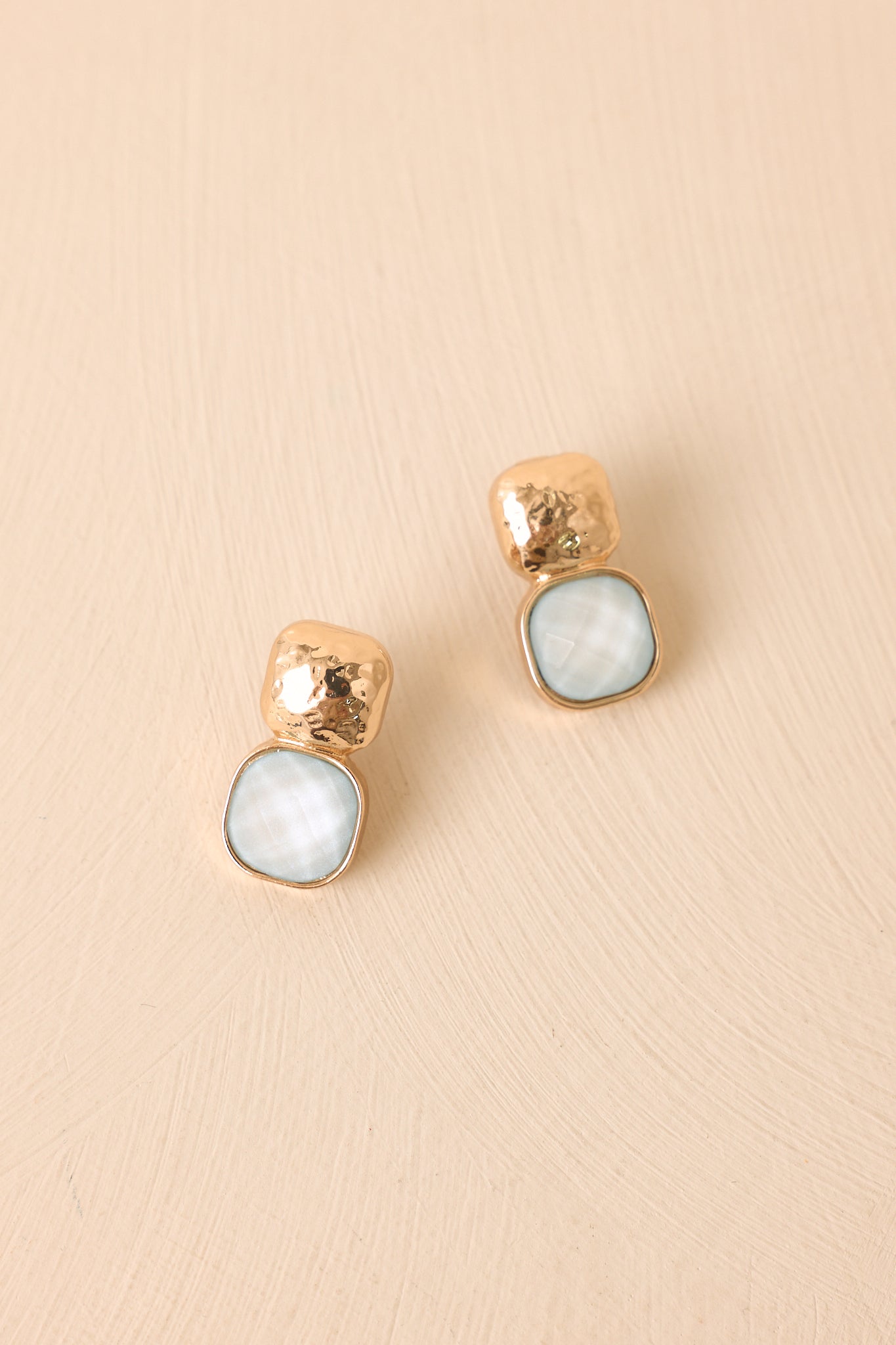 Ice blue cushion cut stone earrings on a beige background featuring textured gold detailing.