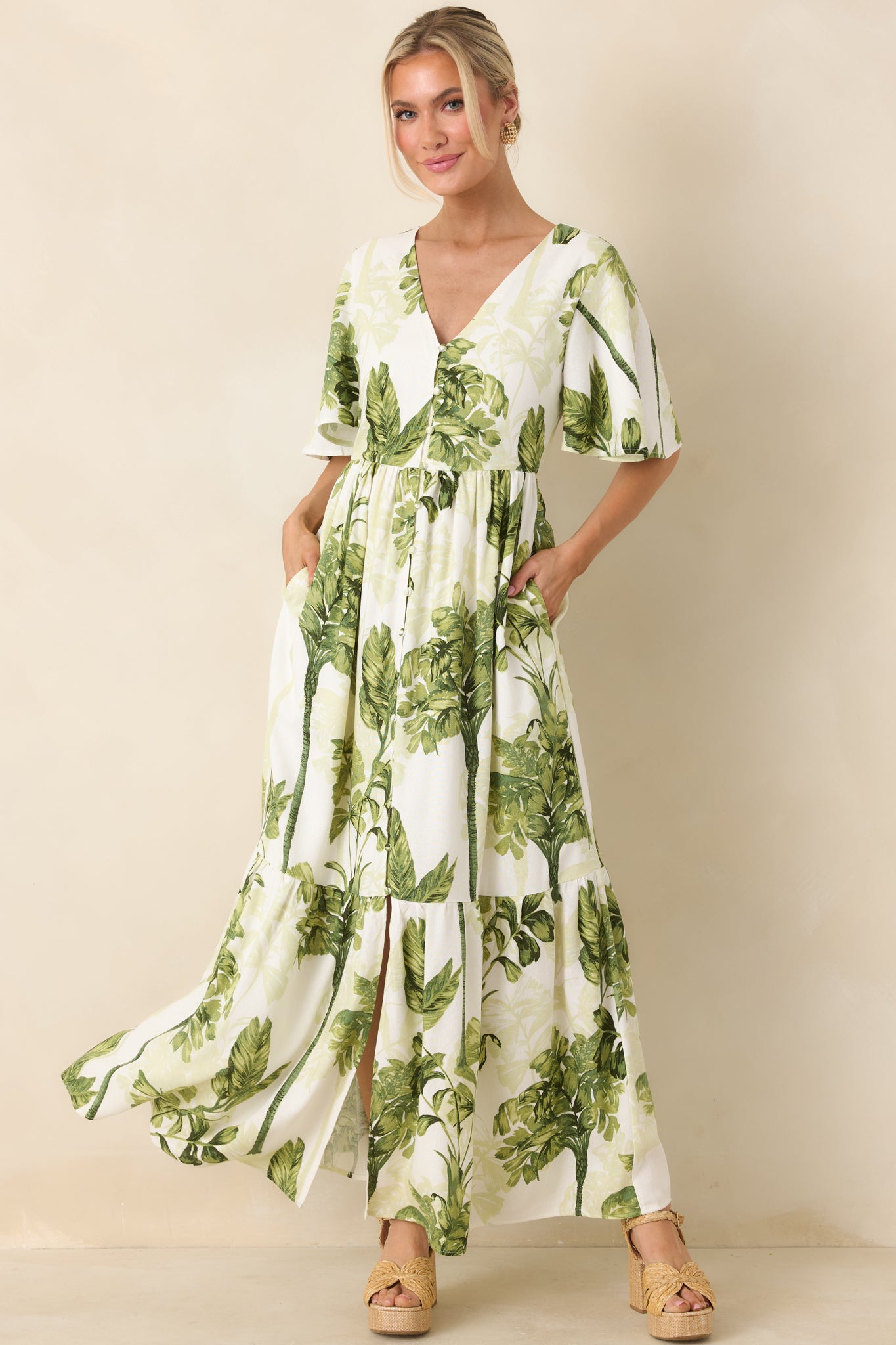 Ivory and green tropical print maxi dress with V-neckline, flutter sleeves, defined waist, and tiered full-length skirt with front button detail.