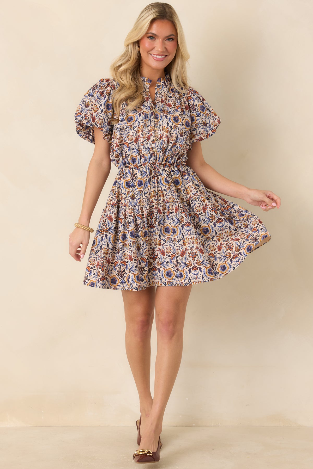 Ivory botanical print mini dress features blue, rust, and golden florals, flutter sleeves, and a tiered skirt.