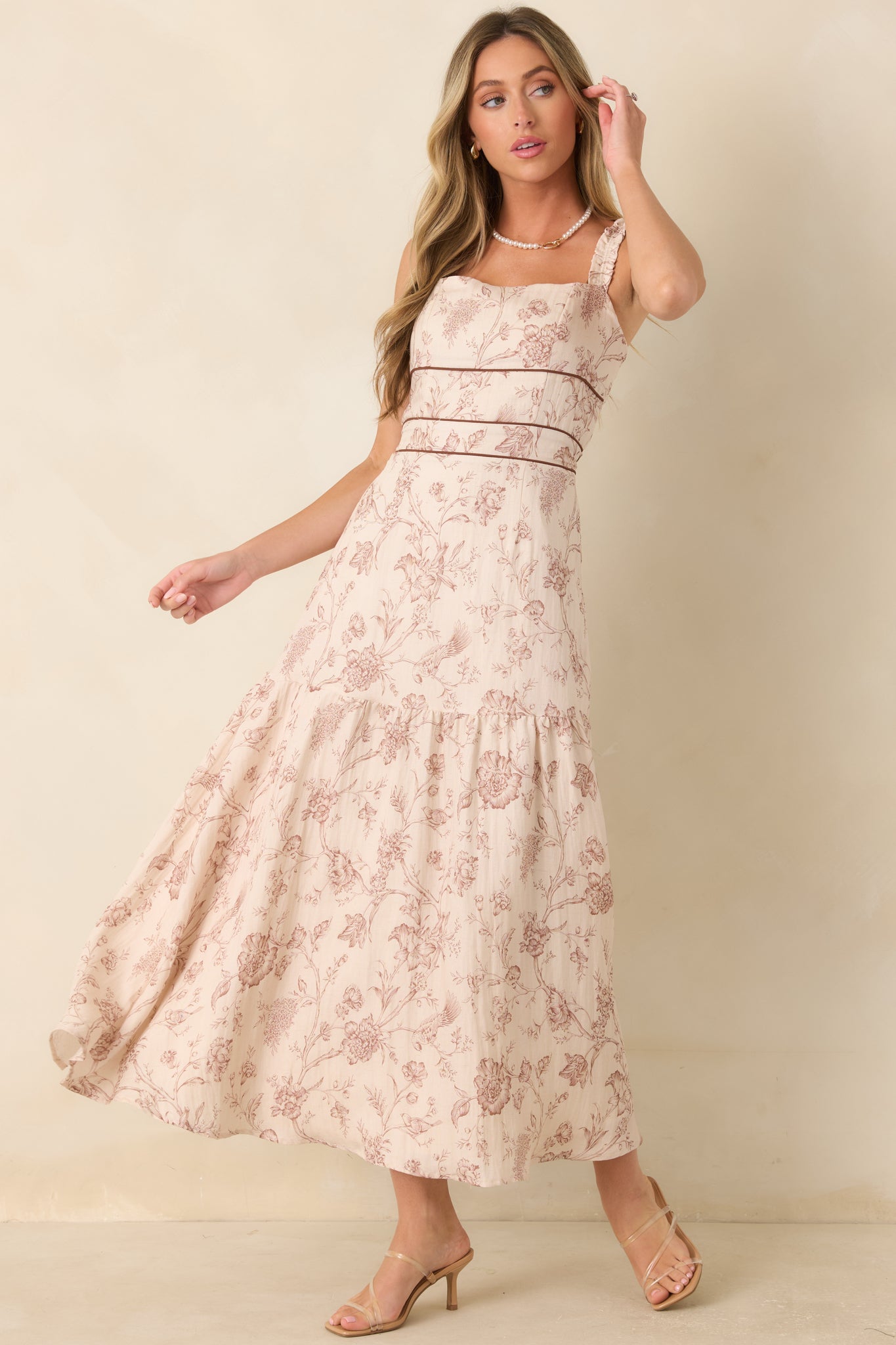 Ivory maxi dress featuring a brown botanical print, ruffled straps, square neckline, contrast piping, and a tiered flowing skirt.