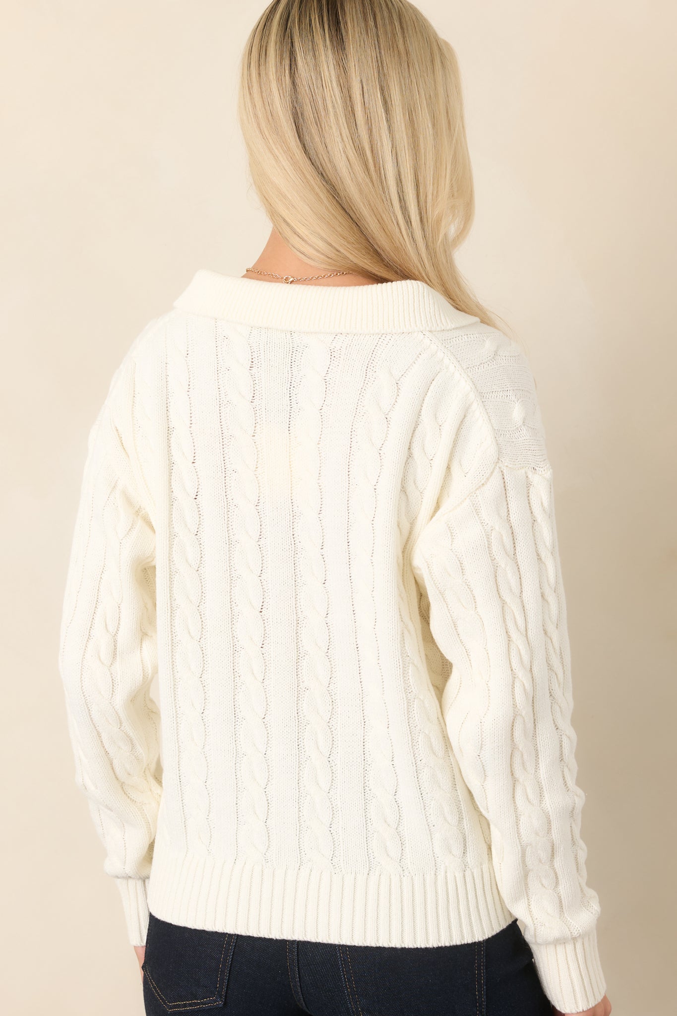Rear view of the ivory sweater highlighting the continuous cable-knit texture, long sleeves, and ribbed hem.