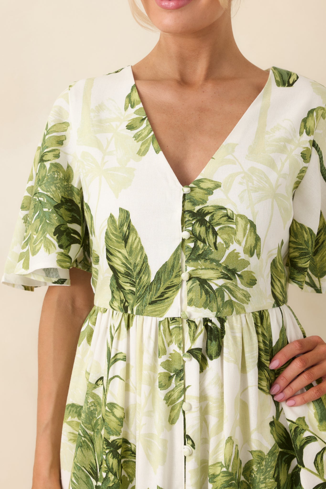 Ivory woven fabric showcasing green tropical print, front button accents, defined waist seam, and softly gathered tiered construction.