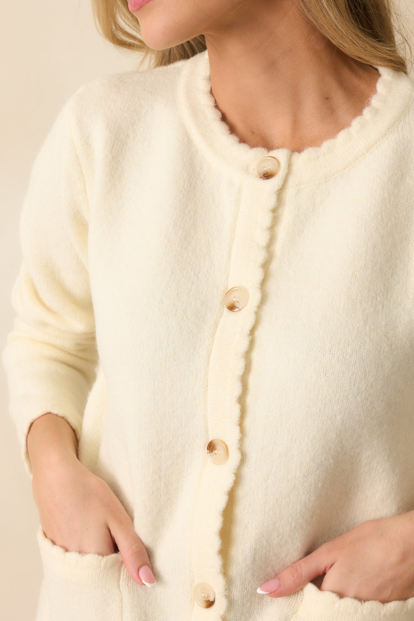 A close-up detail of the ivory cardigan showing a tortoise shell button, scalloped knit edge, and textured fabric.