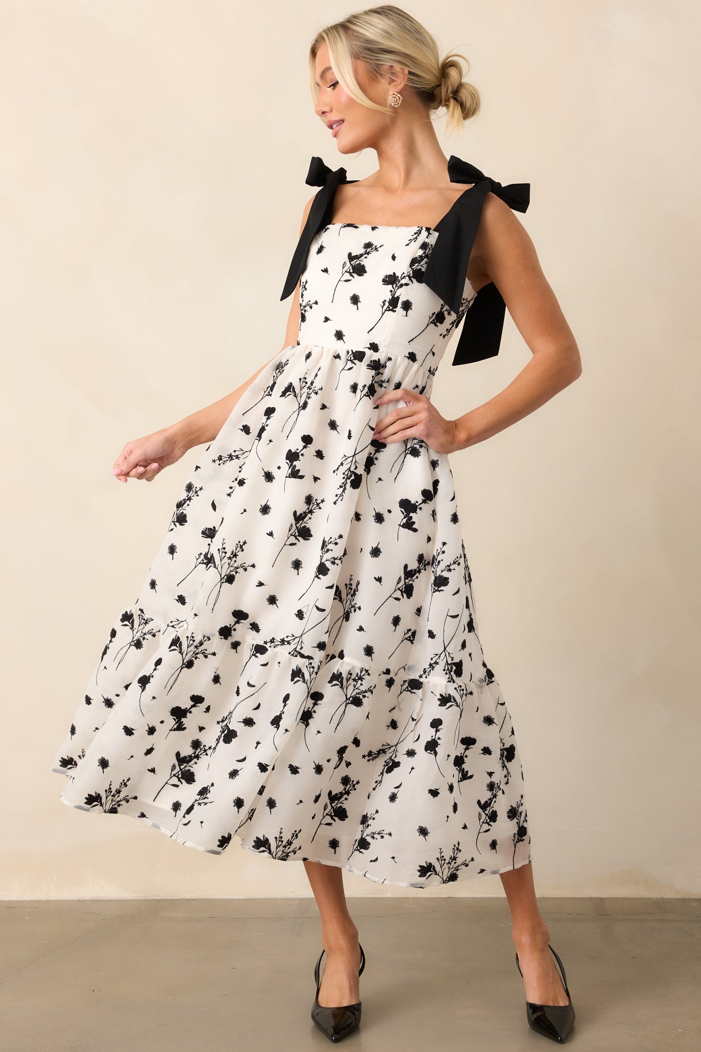 The ivory midi dress, adorned with delicate black floral embroidery, offering a graceful look with adjustable bow straps and a hidden back zipper closure.