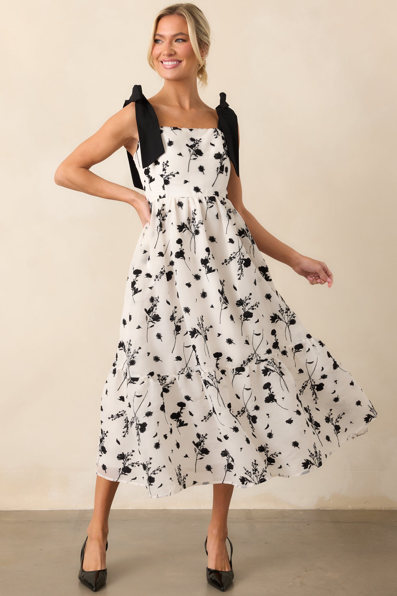 A beautiful midi dress with an ivory base, featuring intricate black floral embroidery and adjustable self-tie bow straps for a custom fit.