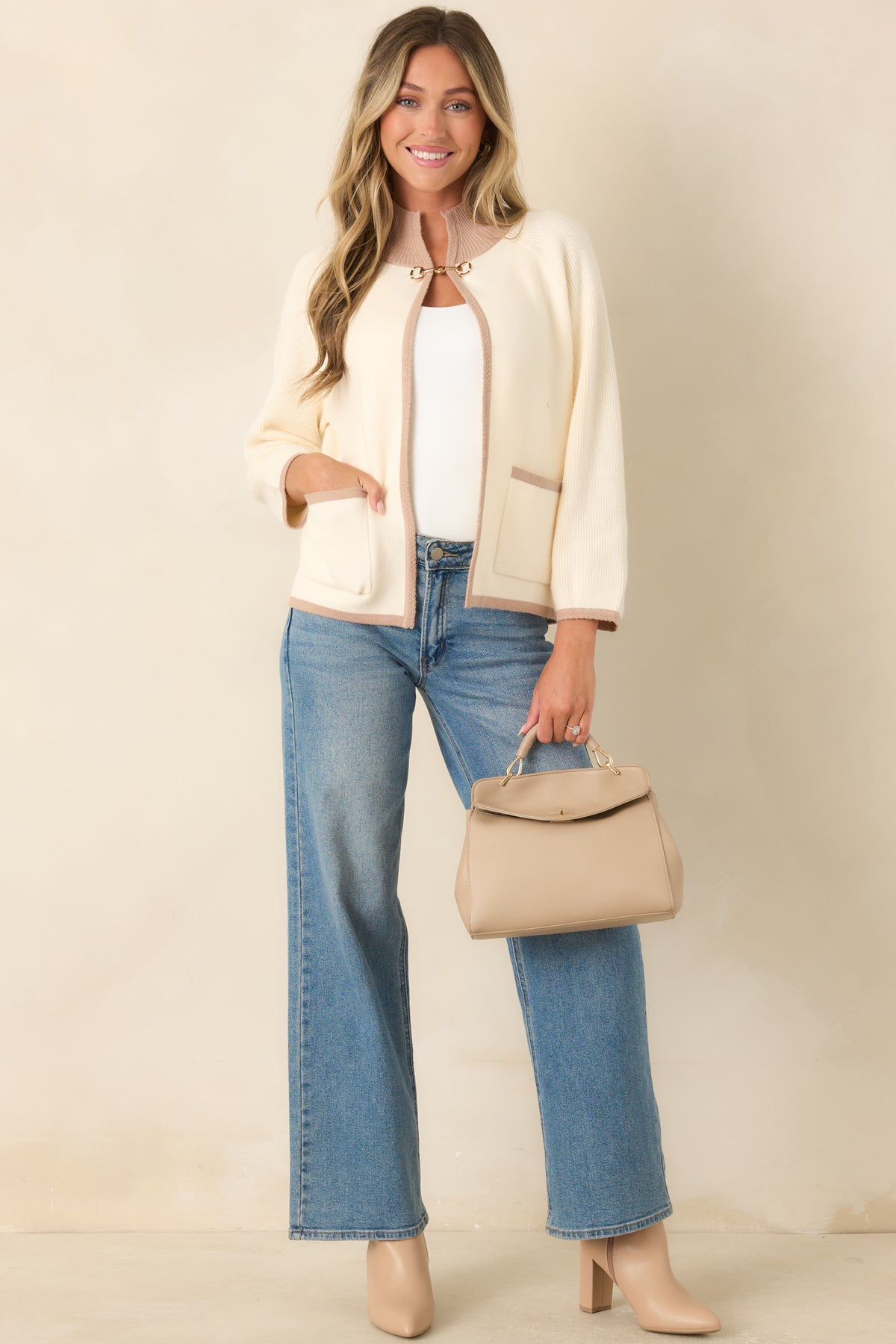 Refined ivory cardigan designed with contrasting taupe edging, front patch pockets, ribbed trim, and a classic equestrian-inspired horsebit closure.