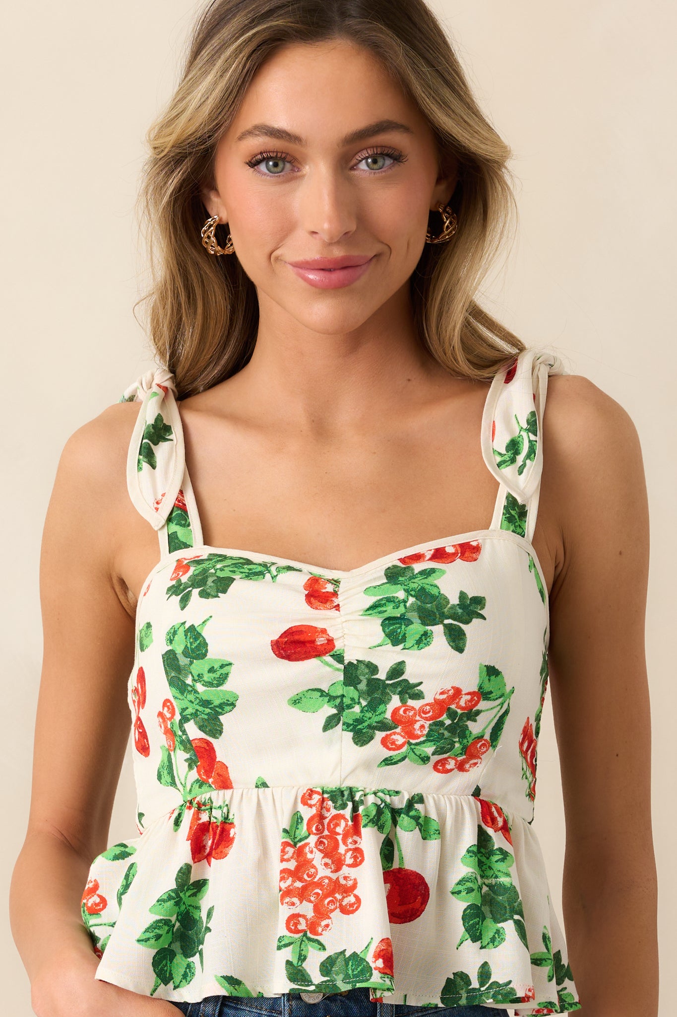 This ivory top features a red and green tomato vine print, a peplum silhouette, and adjustable self-tie straps, offering a playful and feminine look with a cropped length.