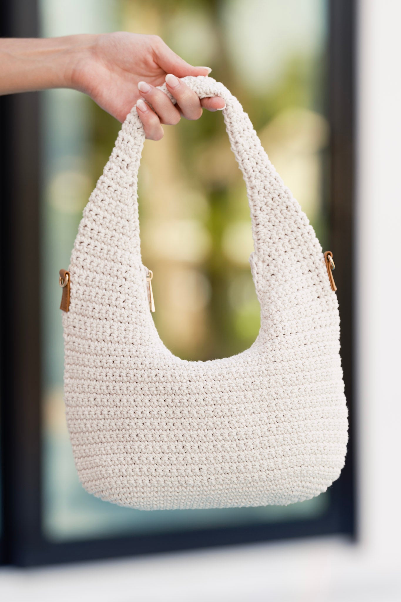 Ivory woven crochet handbag featuring a zipper closure and optional gold crossbody chain, displayed close-up against a blurred outdoor backdrop.