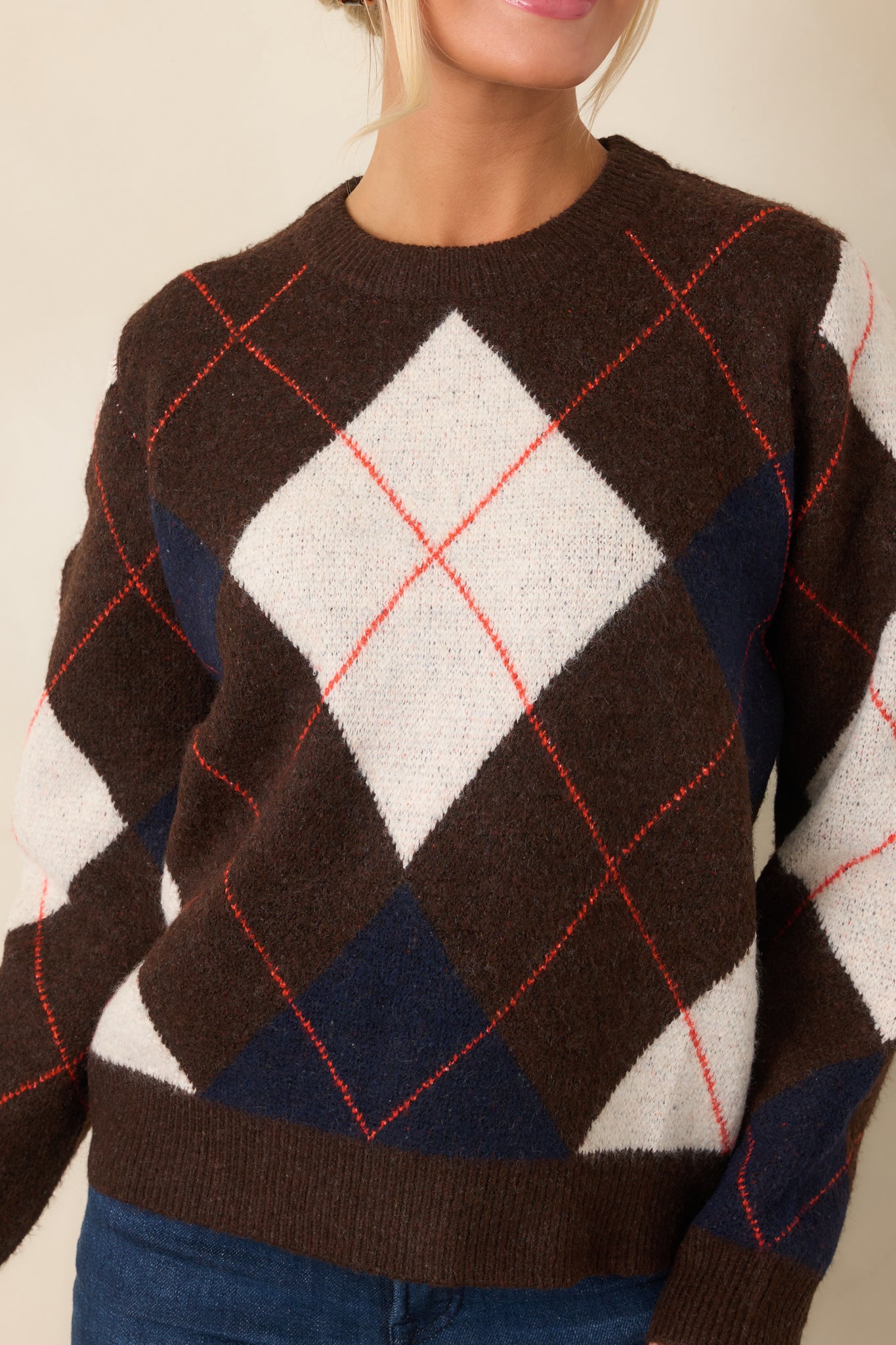 Close-up reveals the soft knit texture, diamond motifs, rust-red lines, and ribbed neckline trim.