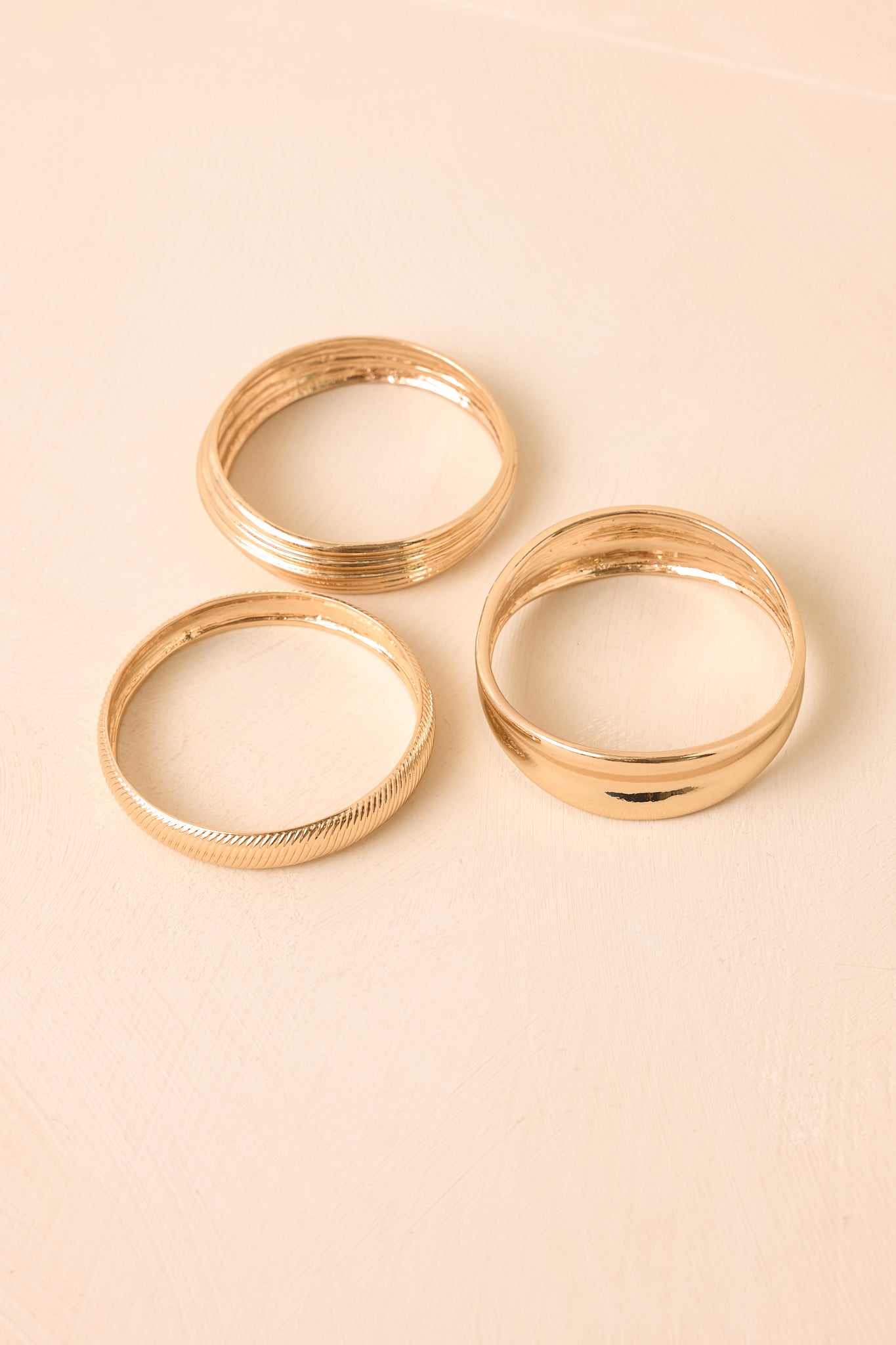 Layered gold bangles with polished, herringbone-inspired, and textured elements presented on a neutral backdrop.