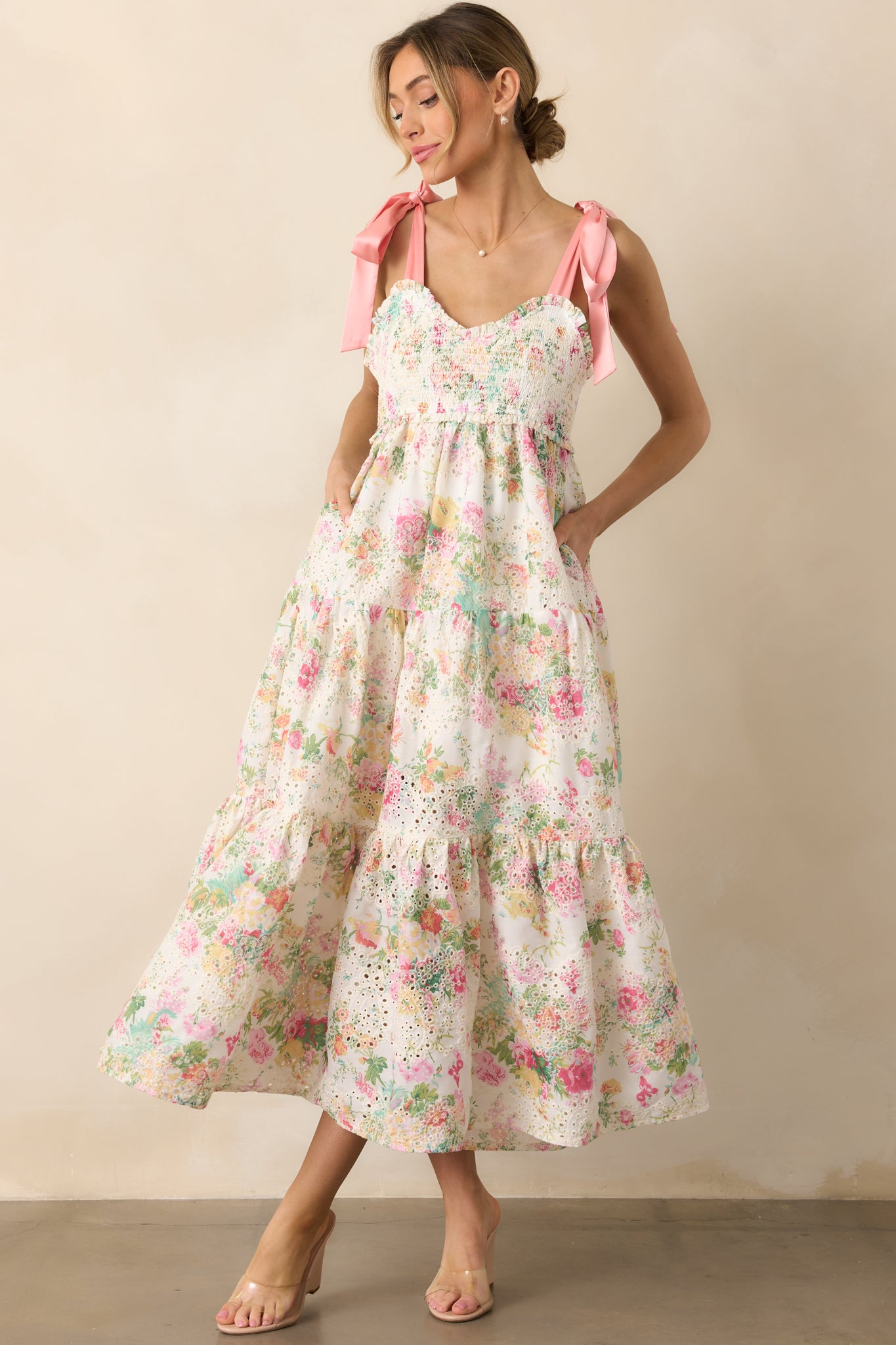 A charming light pink dress with a multicolor floral print, featuring a sweetheart neckline, pink self-tie satin bow straps, and a fully smocked bust, complemented by a tiered skirt and functional pockets.