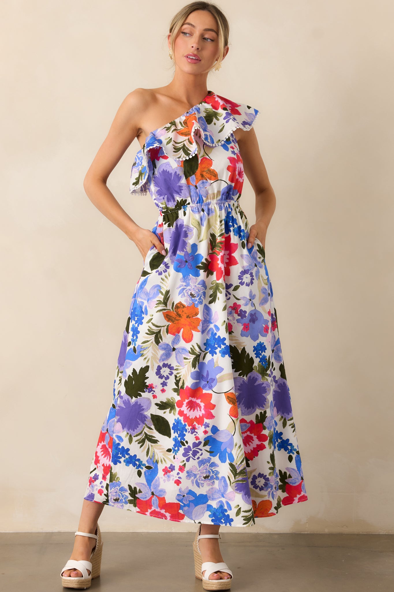 The maxi dress with its striking blue and red floral print, elastic one-shoulder ruffle neckline, and eyelet trim detailing, offering a graceful, feminine silhouette with a functional pocket feature.