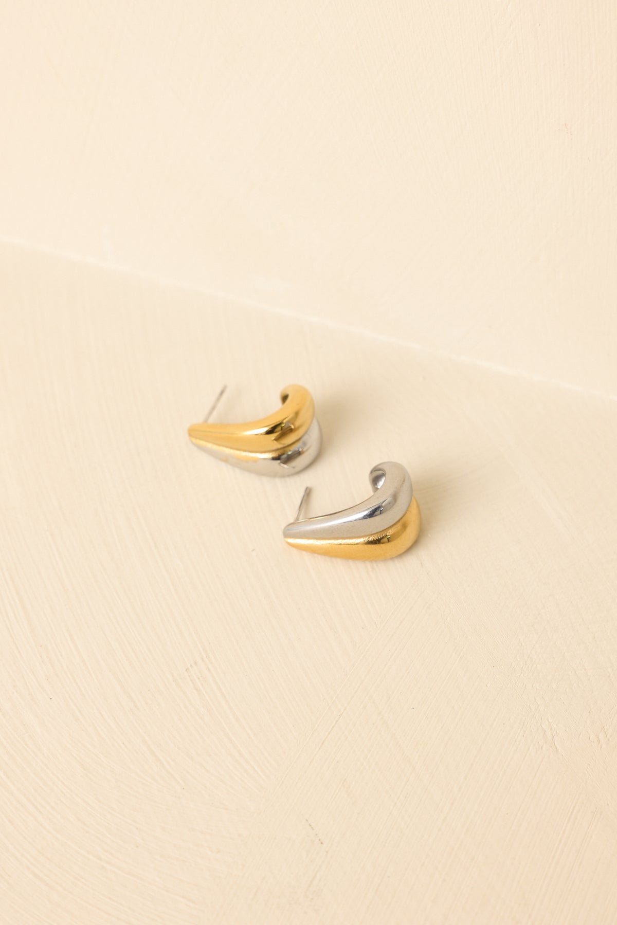 Angled view of mixed-metal sculptural earrings on beige backdrop highlighting shine and dimensional form.