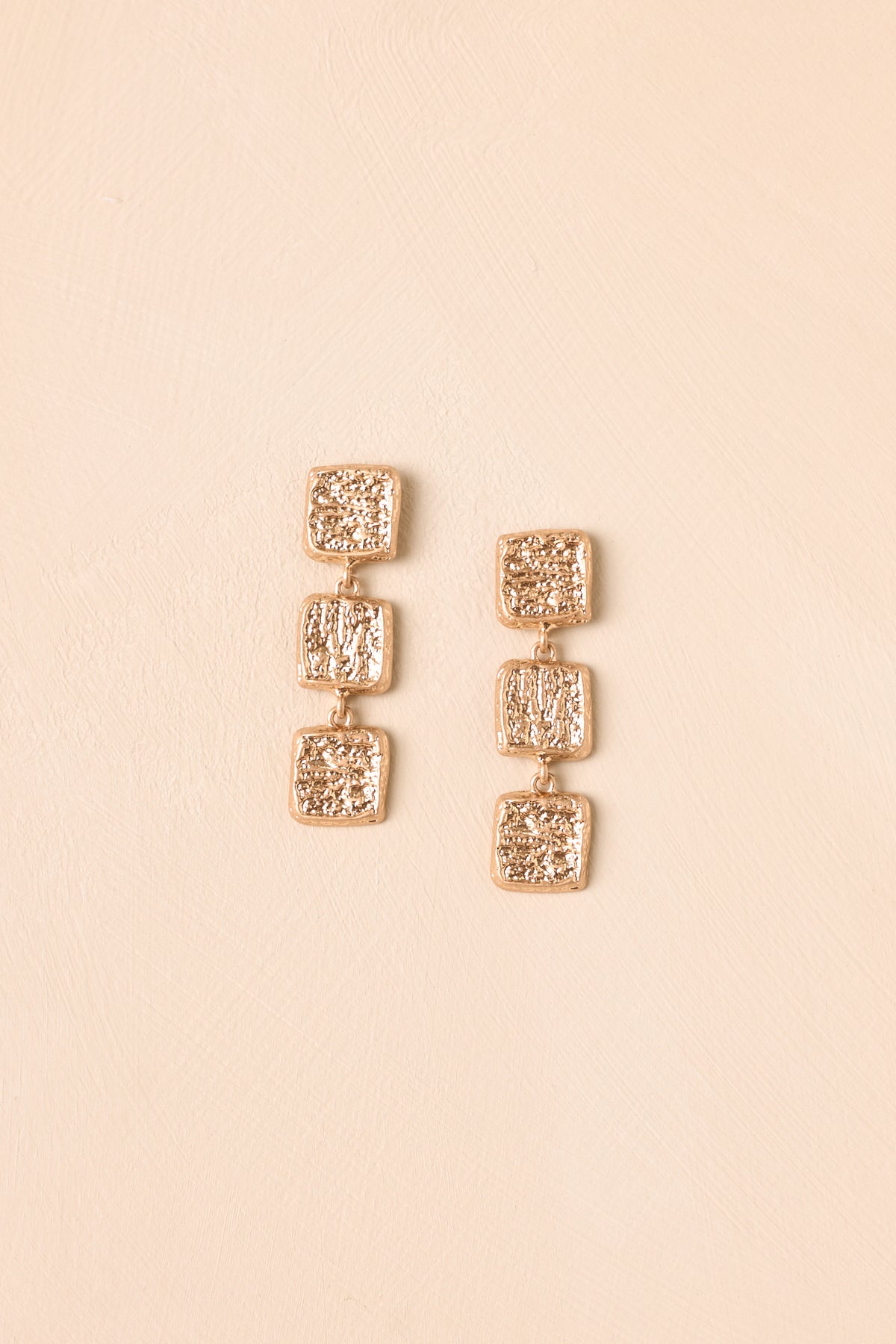 Modern statement earrings presented on a neutral beige backdrop, showcasing layered square details and a softly textured finish.