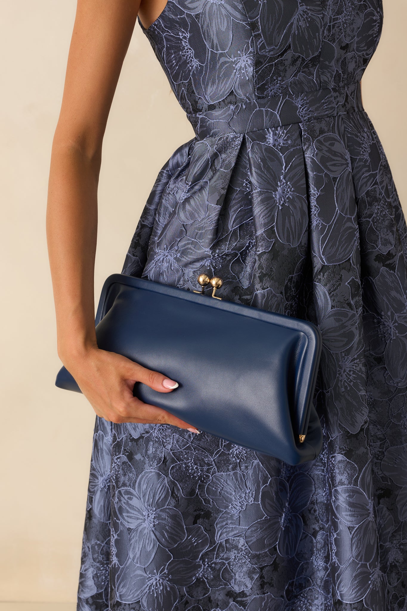 Navy faux leather clutch handbag with a structured frame silhouette and gold-tone kiss-lock closure shown held at the side.
