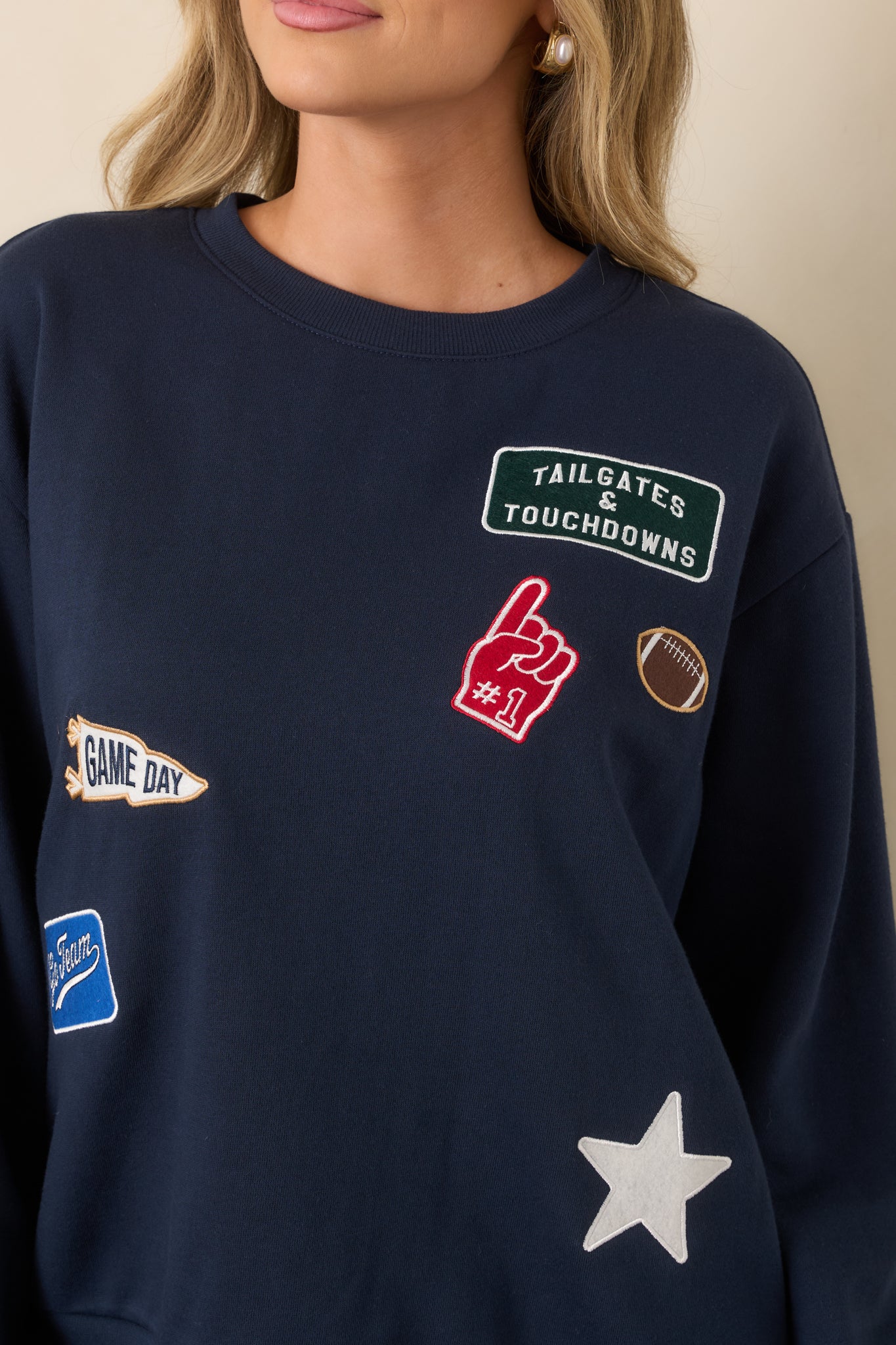Close-up of the navy sweatshirt highlighting its football-themed embroidered and appliqué patches, including a pennant, foam finger, star, and tailgate details.