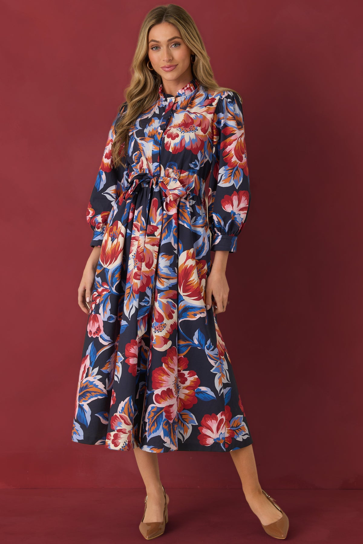 Deep navy cotton midi dress with oversized multicolor florals, a ruffled stand neckline, and billowy sleeves on burgundy background.