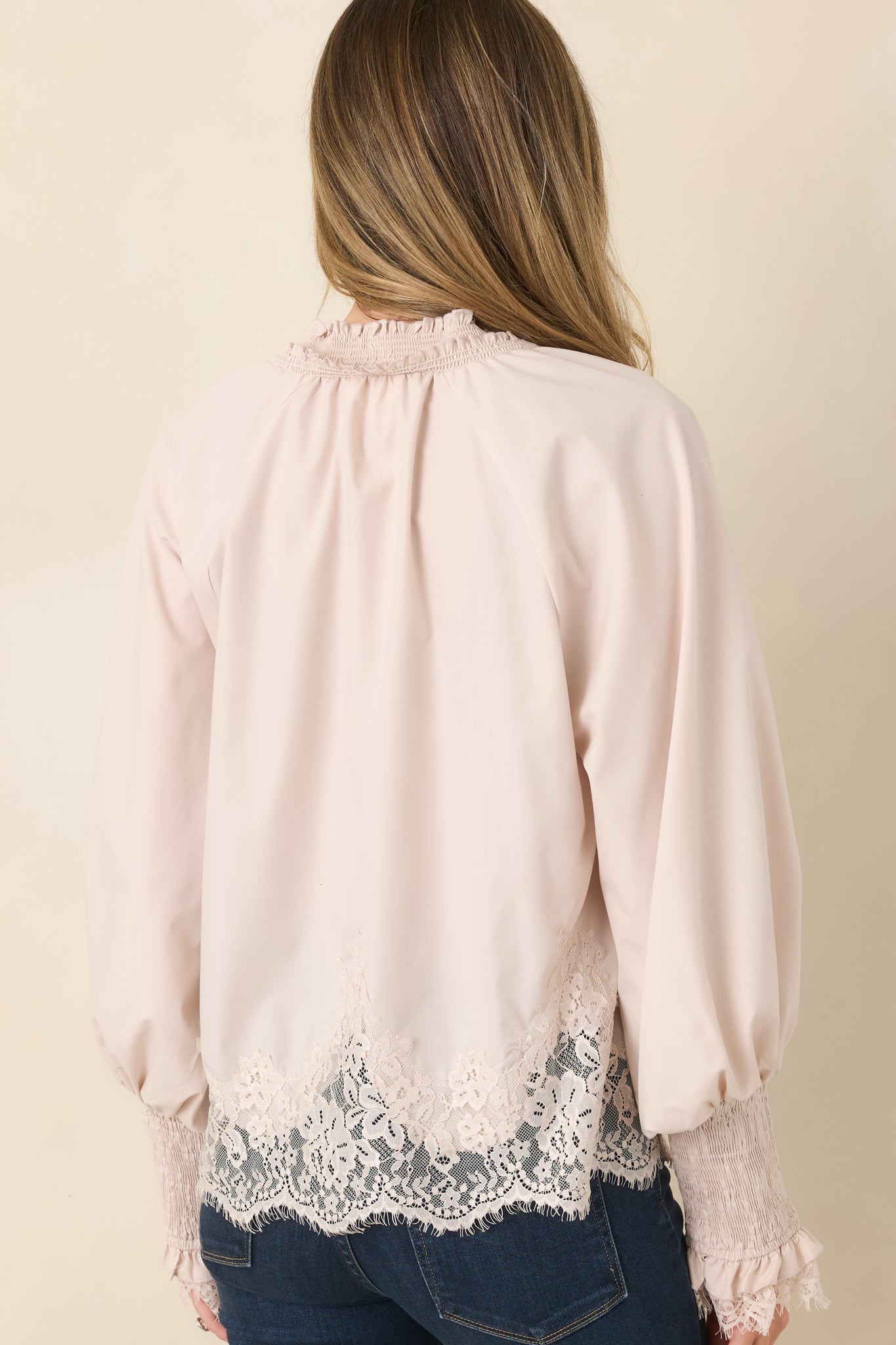 Rear view of the pale neutral blouse displaying the relaxed silhouette, full balloon sleeves, wide smocked cuffs, and floral lace panel extending across the hem.