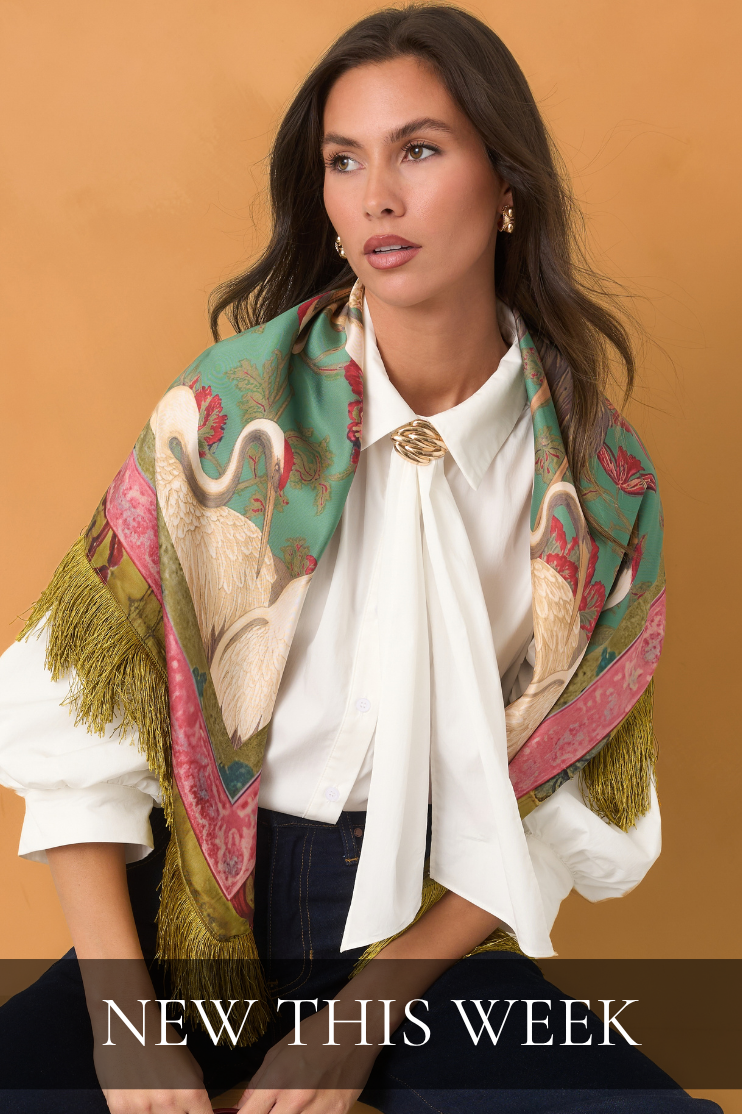 Model wearing a white blouse, patterned scarf with fringe, and dark jeans against an orange background. Text reads ‘New This Week.’