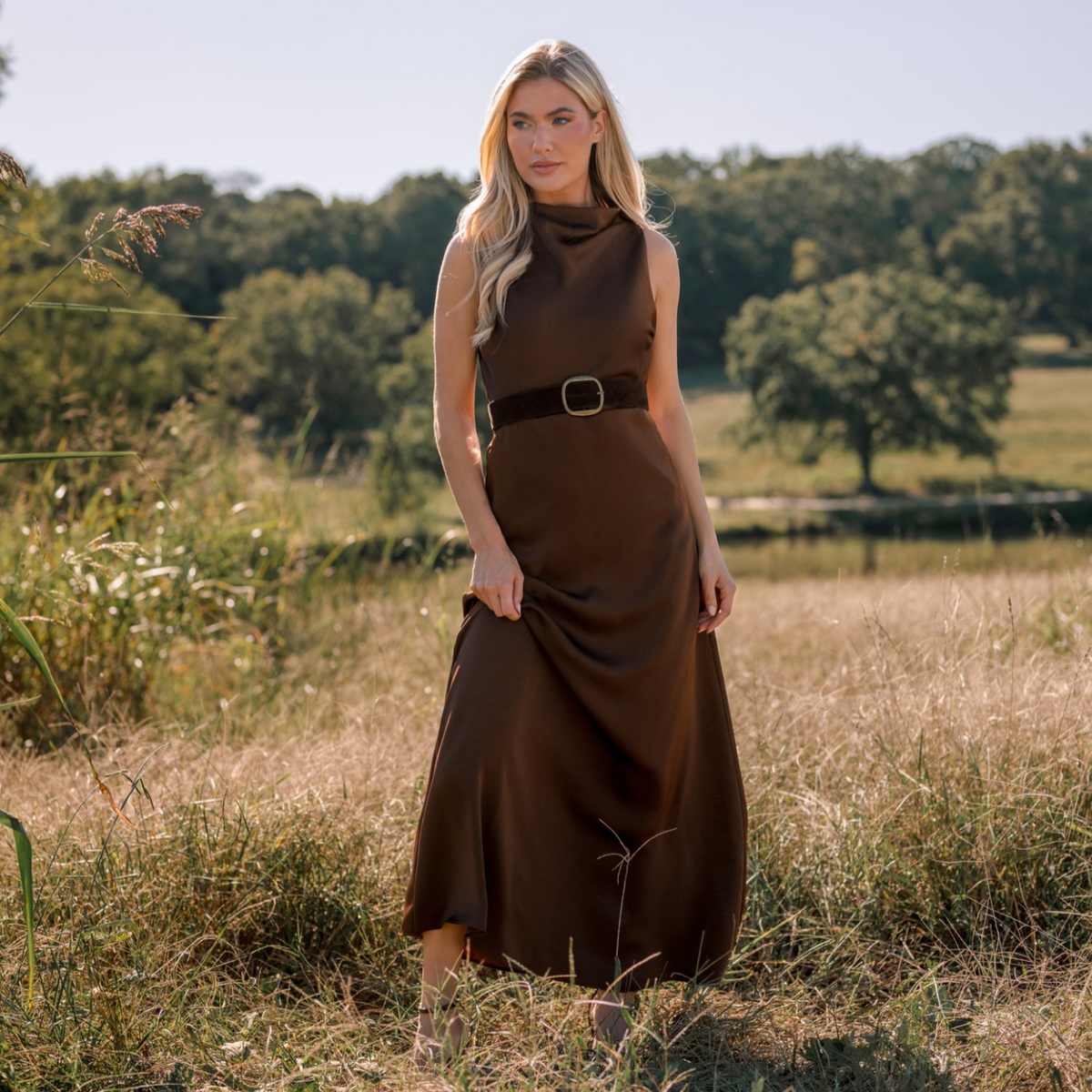 Woman wearing a brown, satin, sleeveless maxi dress in a field. 