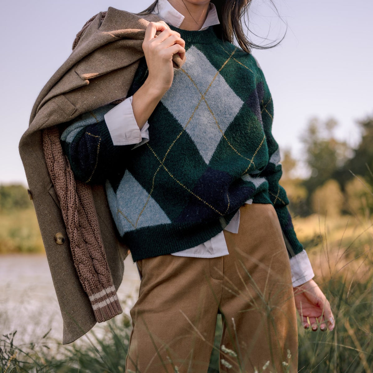 Detail of a green argyle sweater layered over a white shirt with a camel knit-sleeve blazer slung over one shoulder.