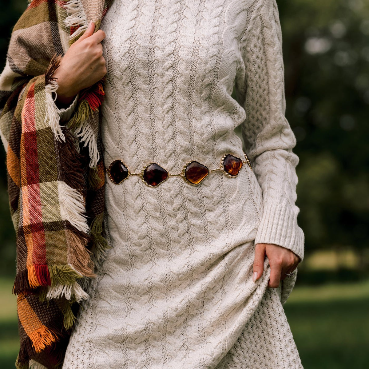 Detail of an ivory cable knit dress with a tortoise link belt and a tartan blanket scarf.