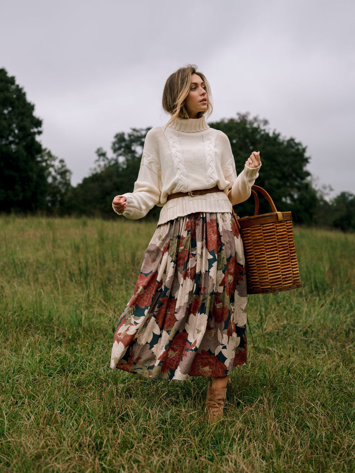 Woman walking through a meadow in a belted ivory cable knit turtleneck and a brown and pink floral tiered midi skirt, carrying a wicker basket.