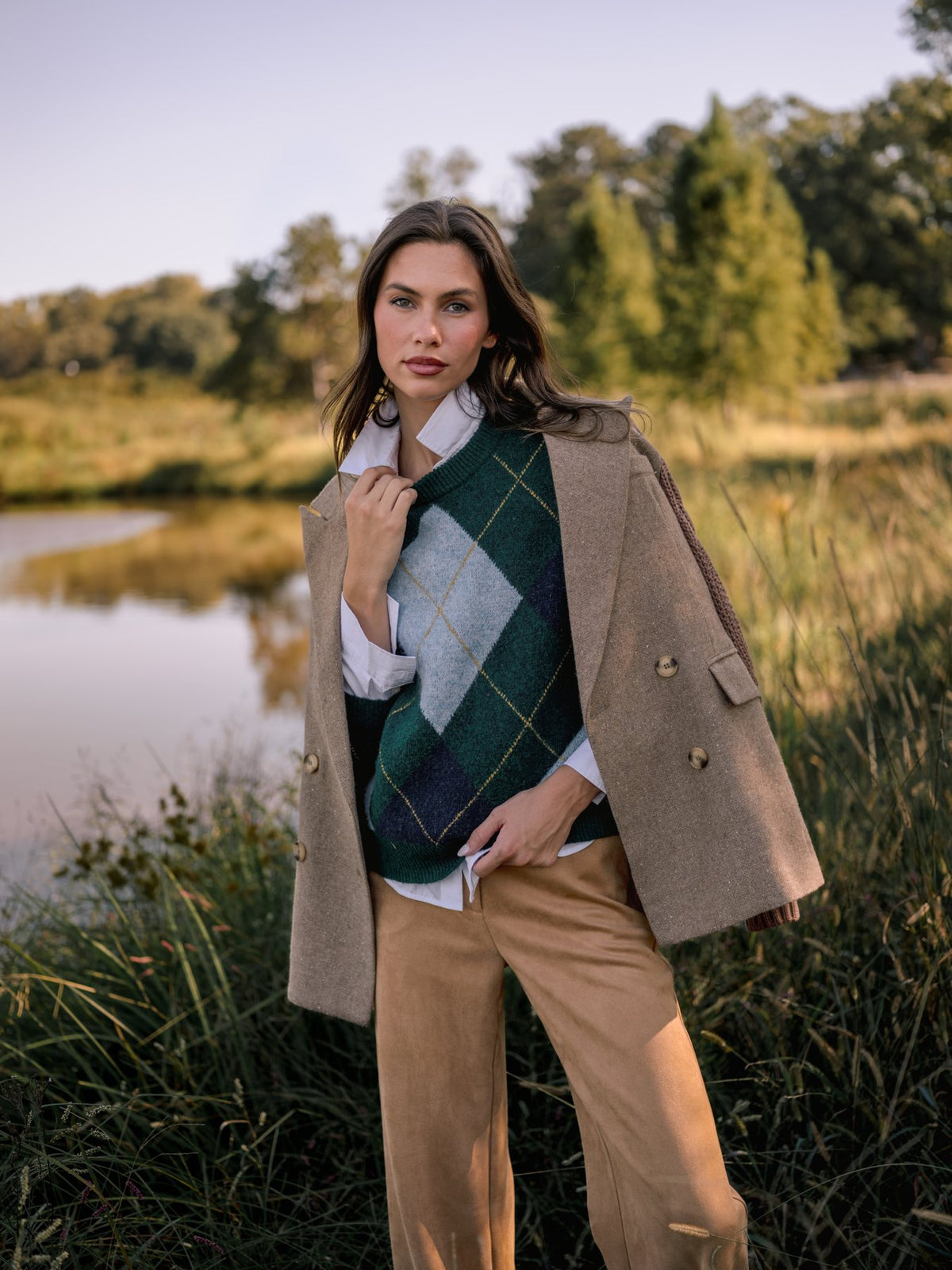 Old money style outfit: hunter green argyle crewneck sweater over a white collared shirt, camel wool blazer draped over the shoulders, and camel trousers beside a pond.