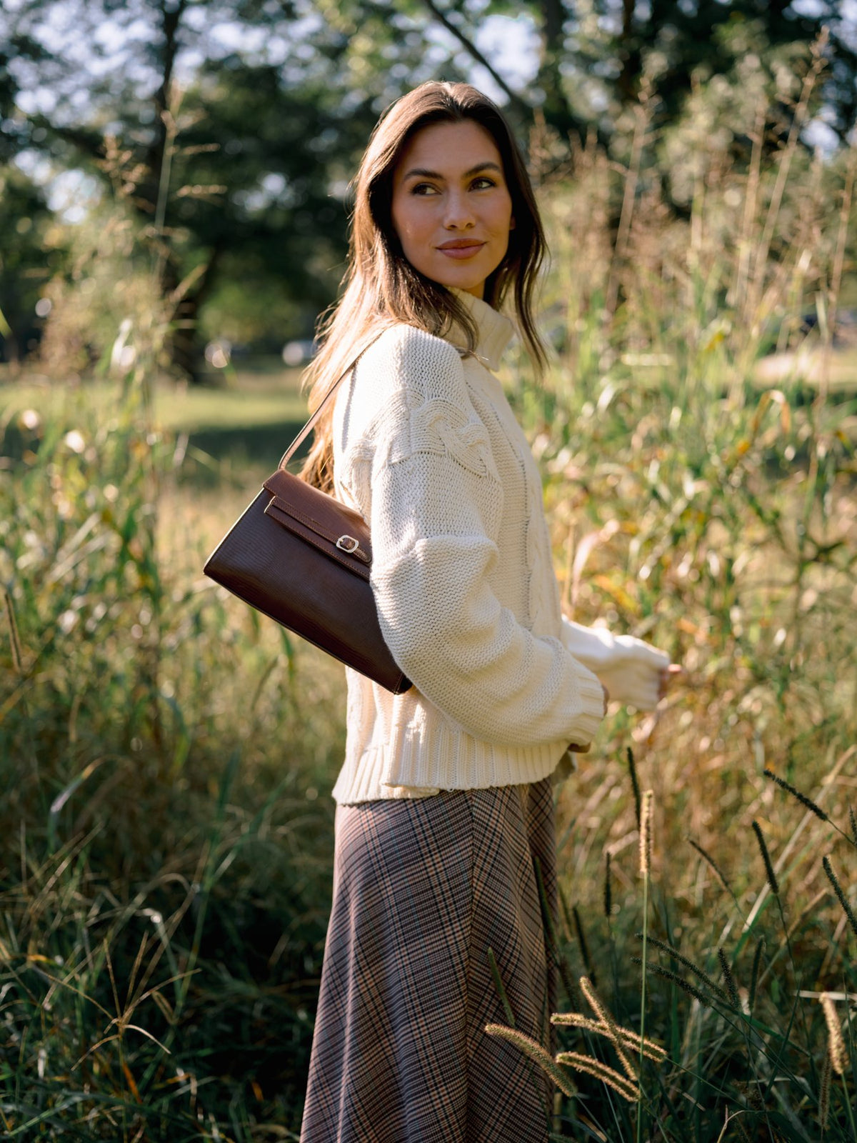Woman in an ivory cable knit turtleneck and brown plaid midi skirt carrying a brown structured handbag through tall grass.